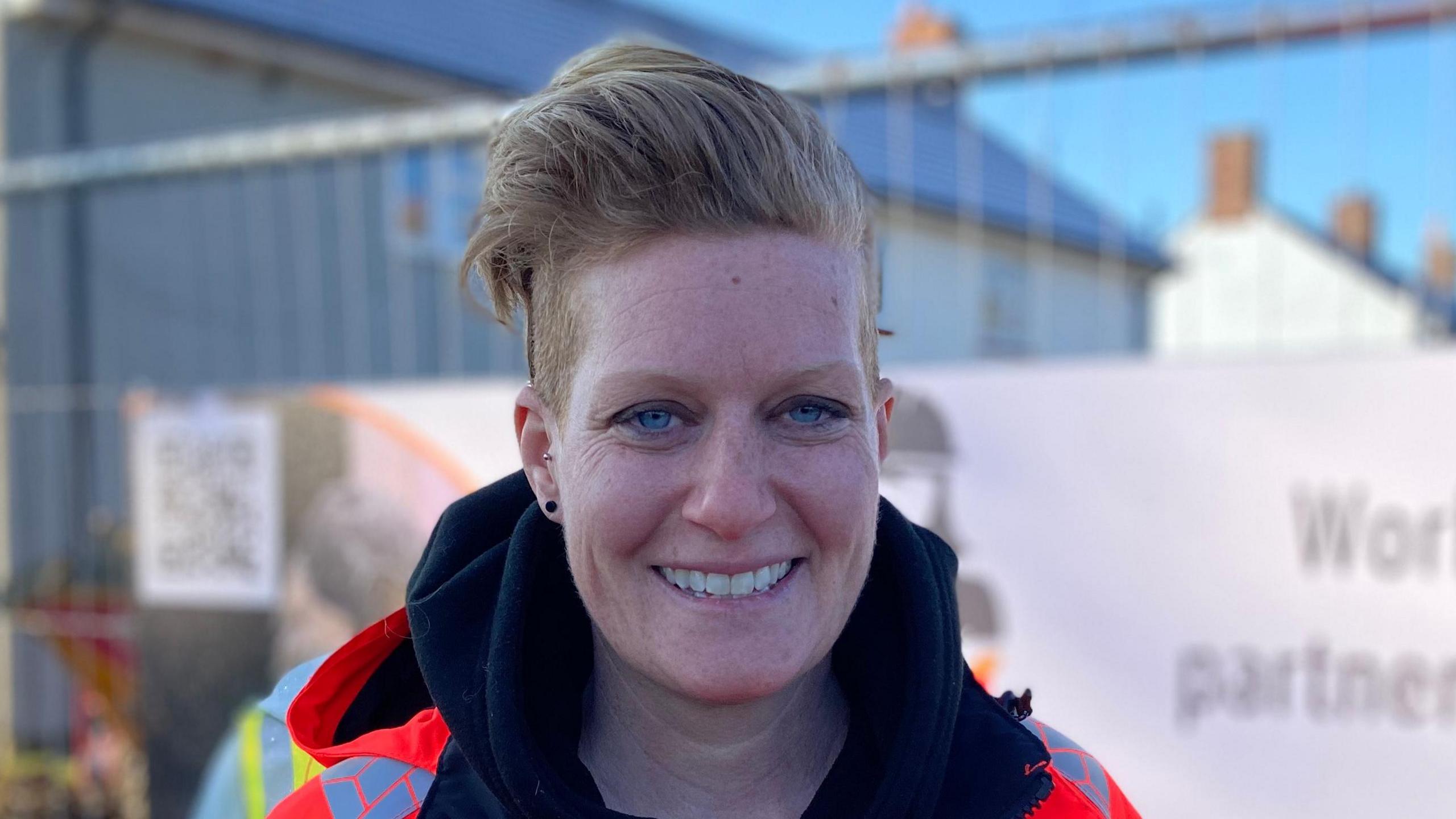 Kat Parsons smiles at the camera in front of a construction site. She has short blonde hair and wears an orange hi-vis jacket with a black hoodie underneath.