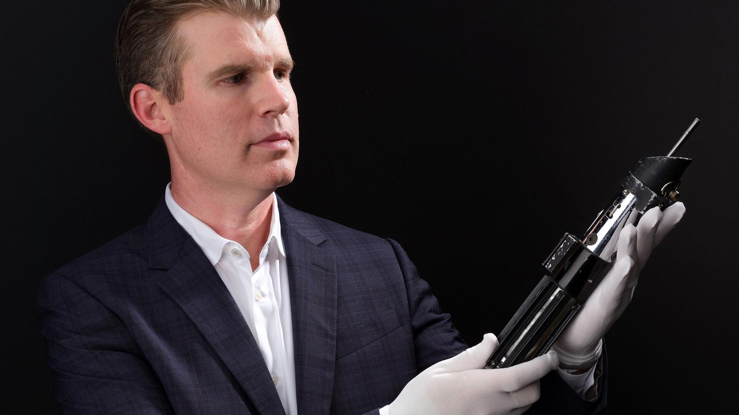 A man in a suit and wearing white gloves is carefully holding and admiring a lightsaber prop against a black background