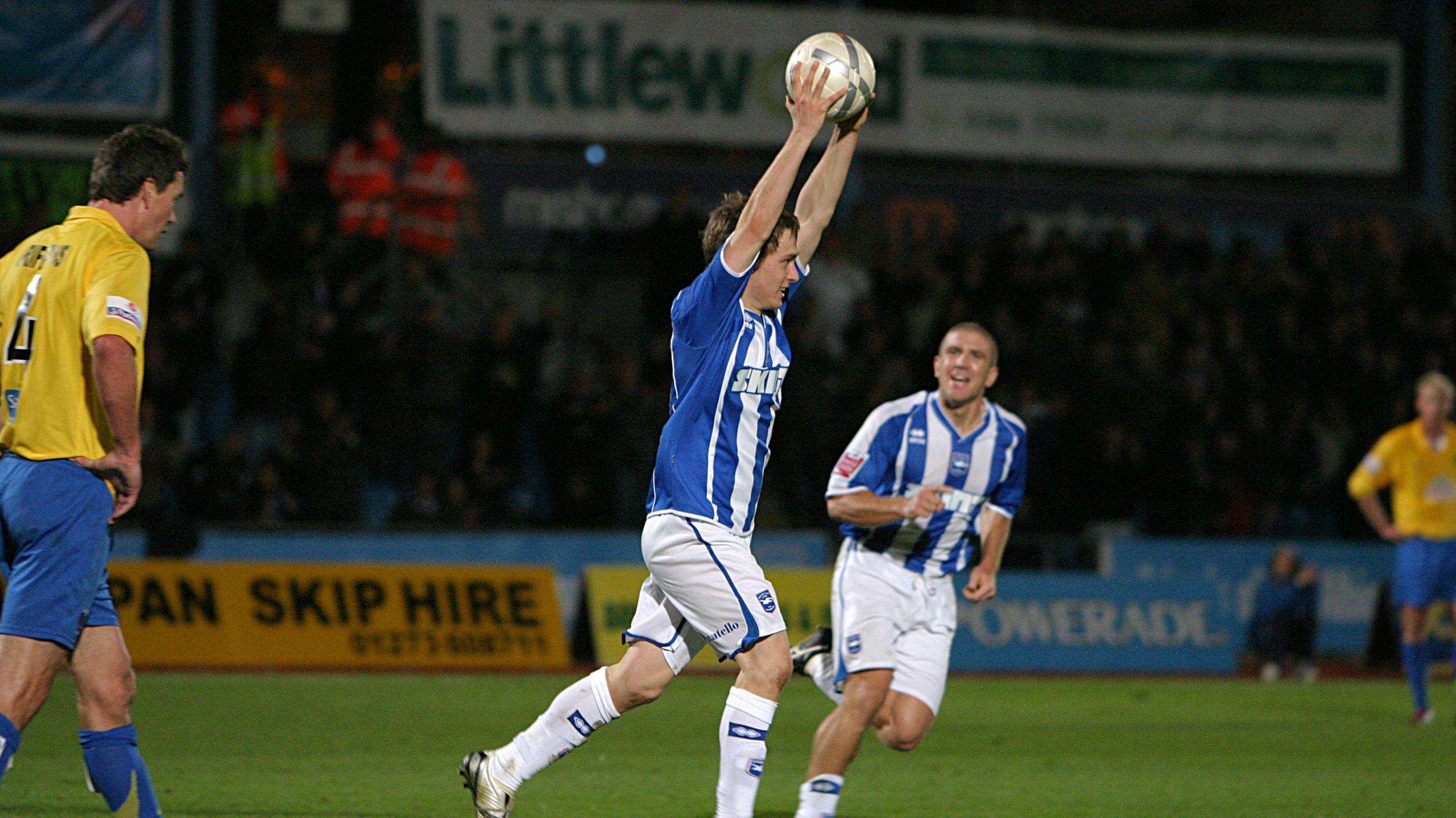 Jake in 2006 running and holding the ball above his head after scoring three goals. He is wearing a blue and white striped kit with white shorts and socks. The opposition are wearing yellow and walking away and there is a small stand of the Withdean stadium behind him with yellow and blue advertising hoardings between the pitch and stand