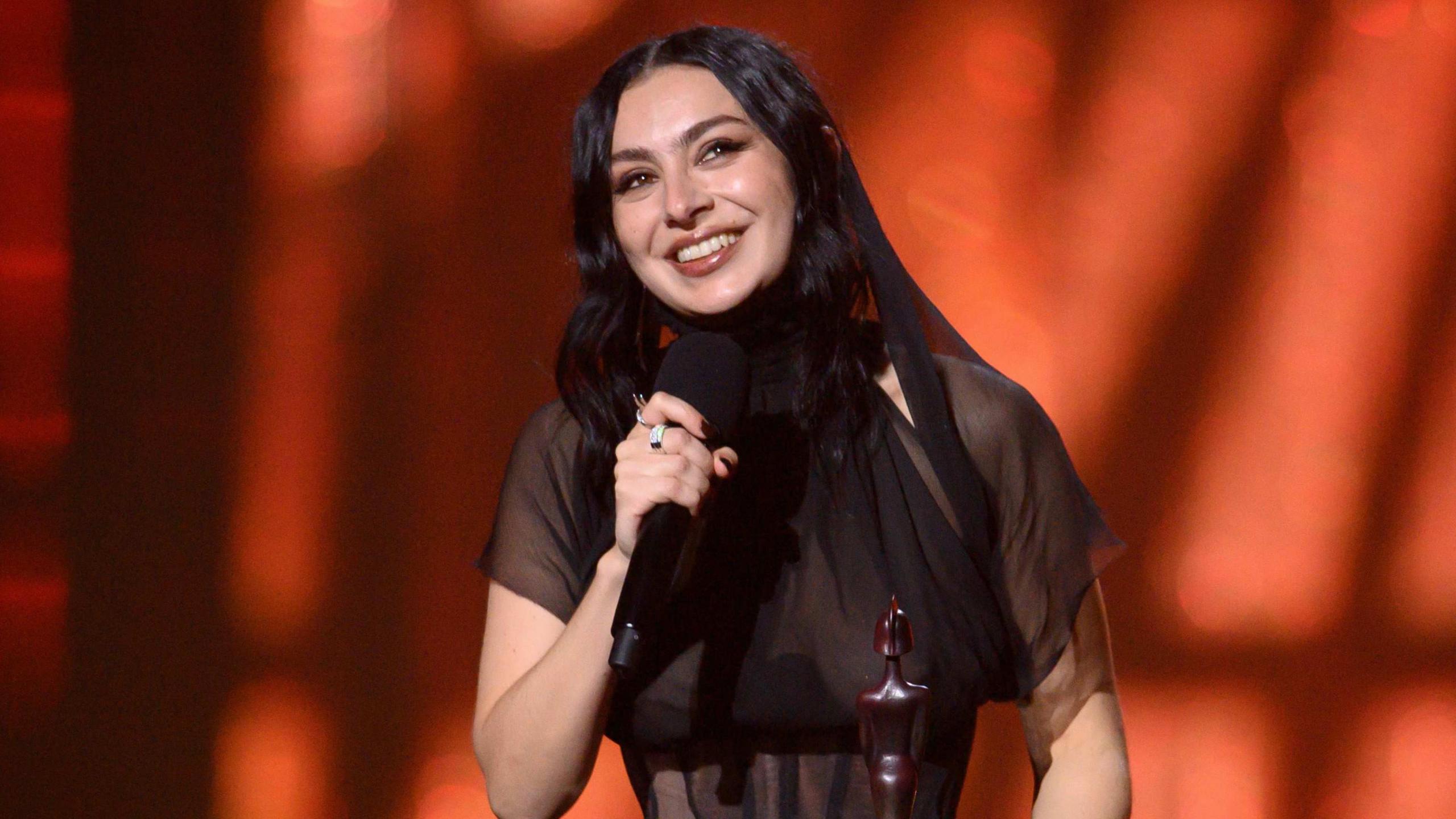 Charli XCX smiling while accepting one of her five Brit awards on stage last year. She is wearing a sheer black dress.