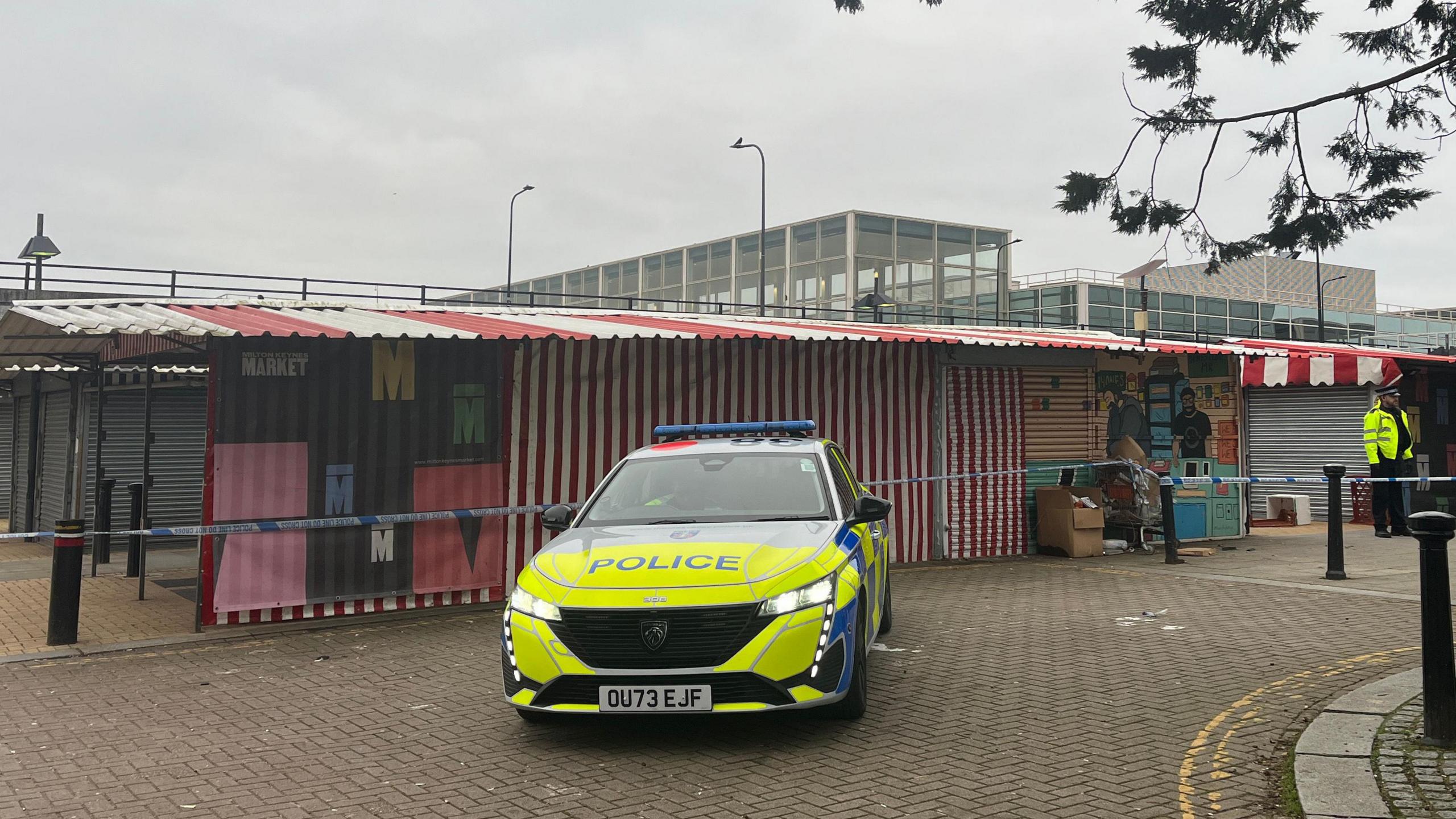 Market stalls with a red and white striped canopy can be seen within a blue and white taped police cordon. A police car sits in front of the stalls and a police officer is stood by the side of the cordon. 