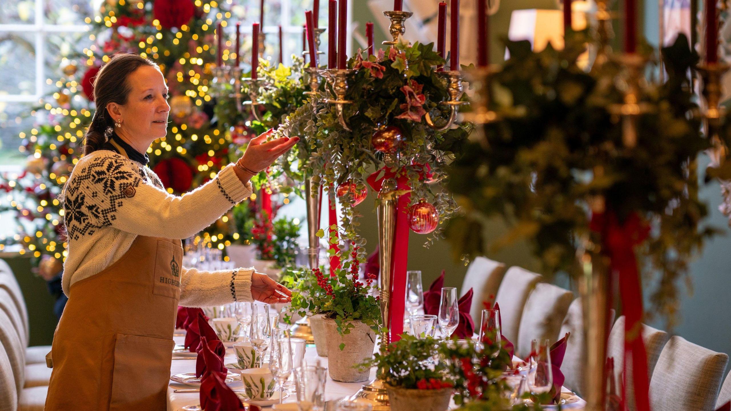 A woman in a festive jumper and brown apron prepares table decorations at Highgrove, the King's estate in Gloucestershire. She is standing next to a long table with ornate decorations centred around tall red candles, and a large Christmas tree is visible in the background