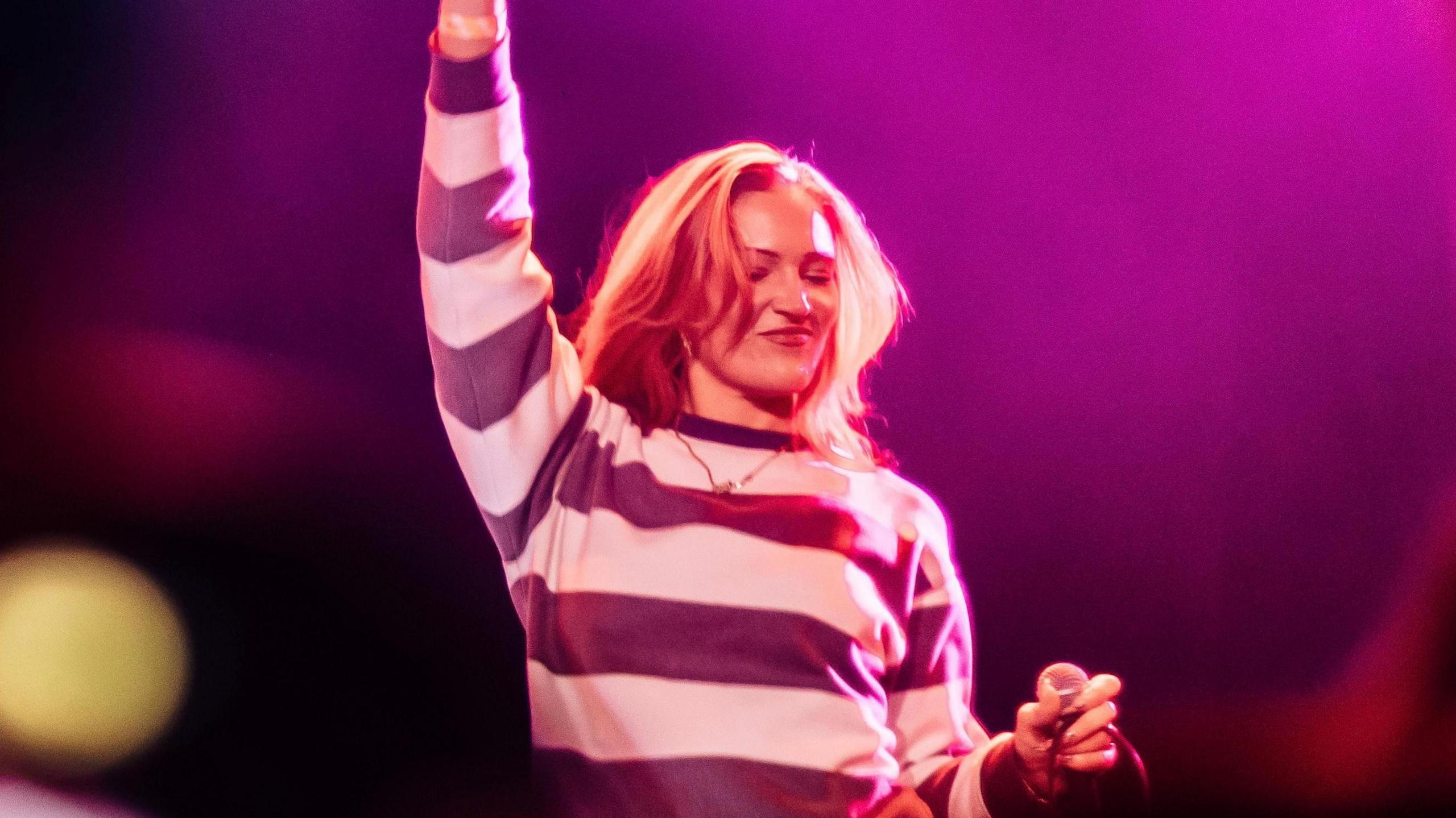 Anna is standing on stage with purple lighting surrounding her. She is raising her hand and smiling while holding a microphone.