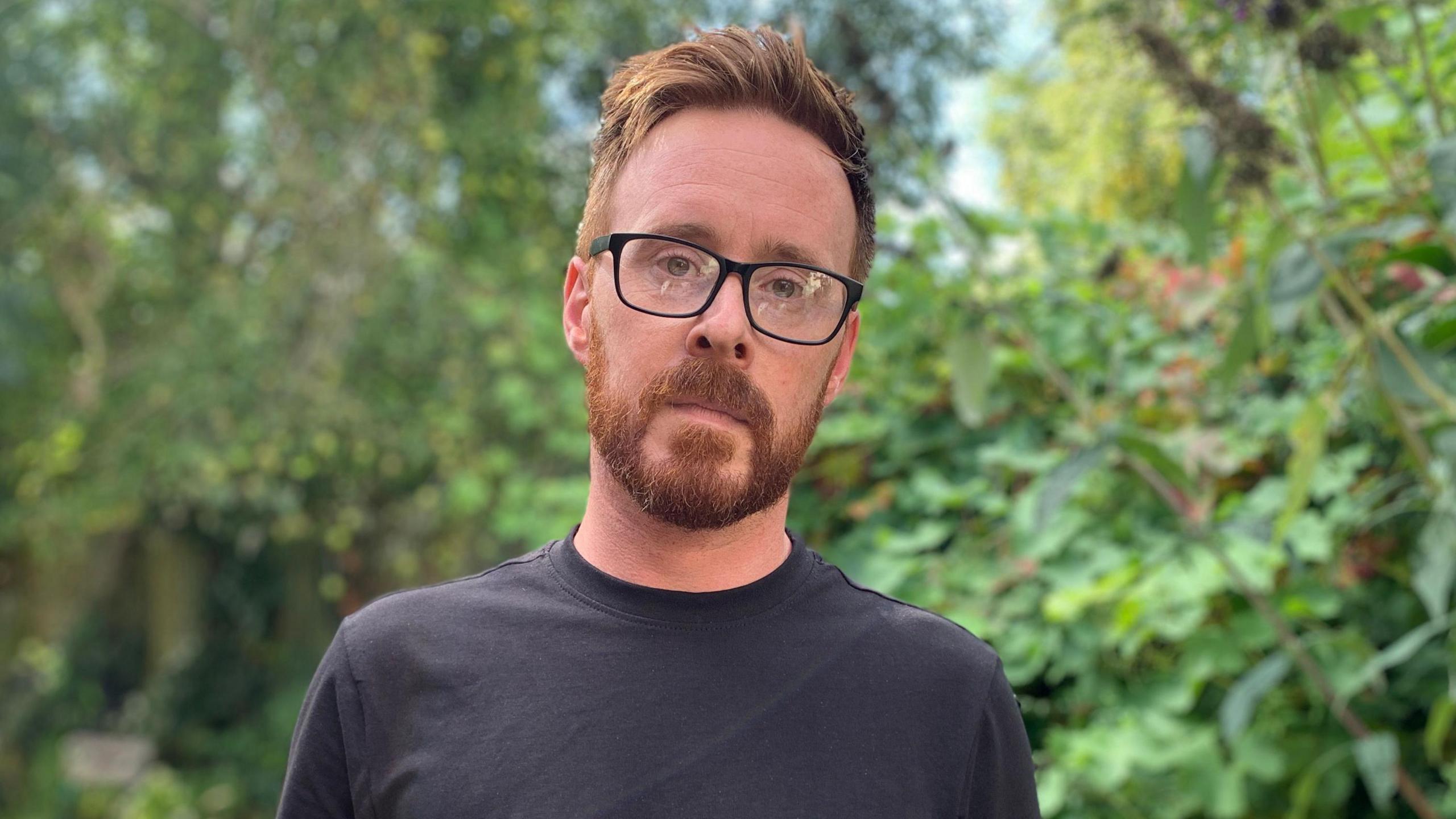 Stephen Webb wearing glasses and a black T-shirt. He is standing outside, and there are trees and bushes behind him