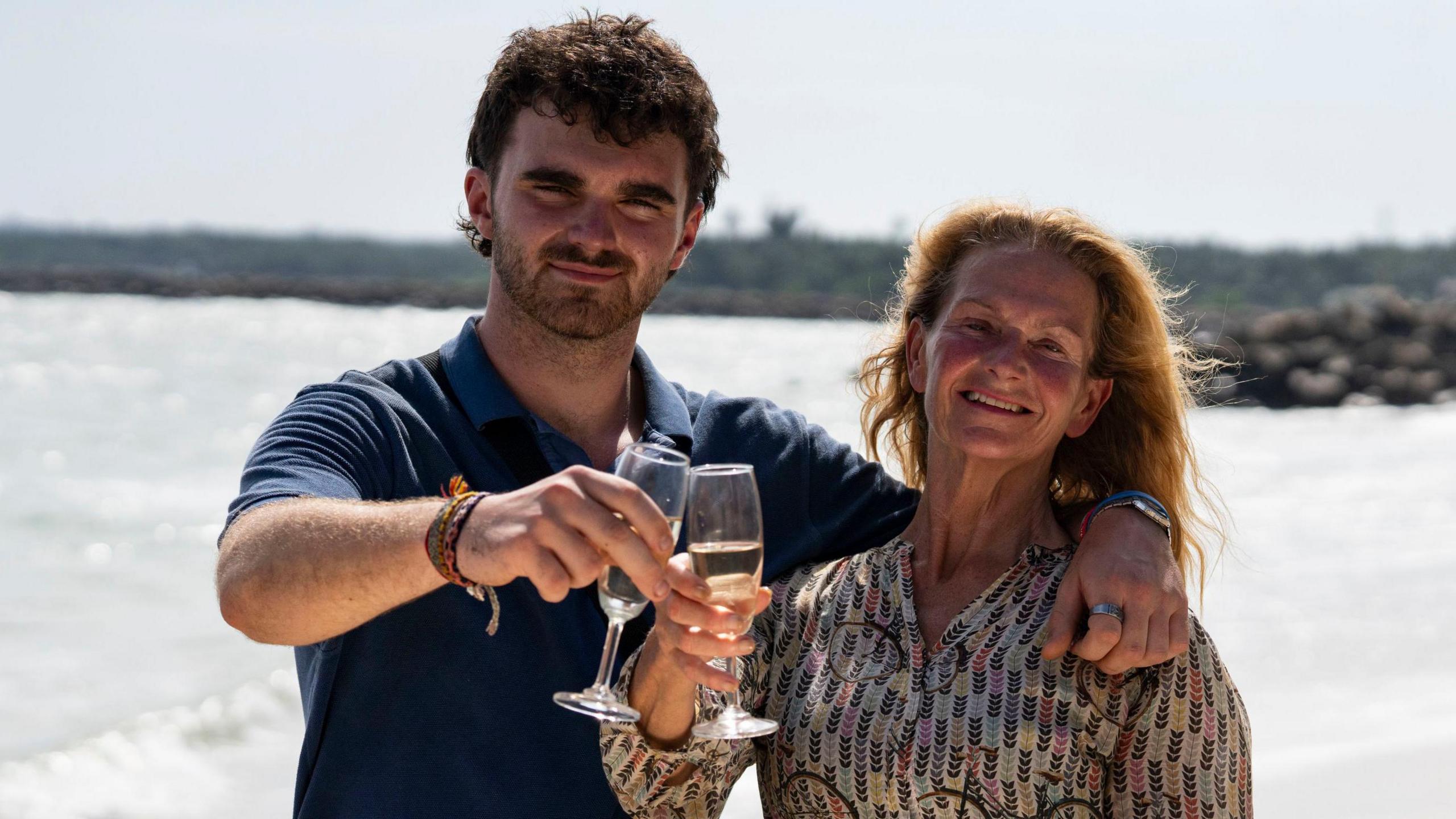 Tom and Caroline Bridge standing outside on a beach both holding a glass. Tom is on the left and is smiling and looking at the camera and is wearing a navy T-shirt. While Caroline, is standing next to Tom on the left, she is looking and smiling directly at the camera.