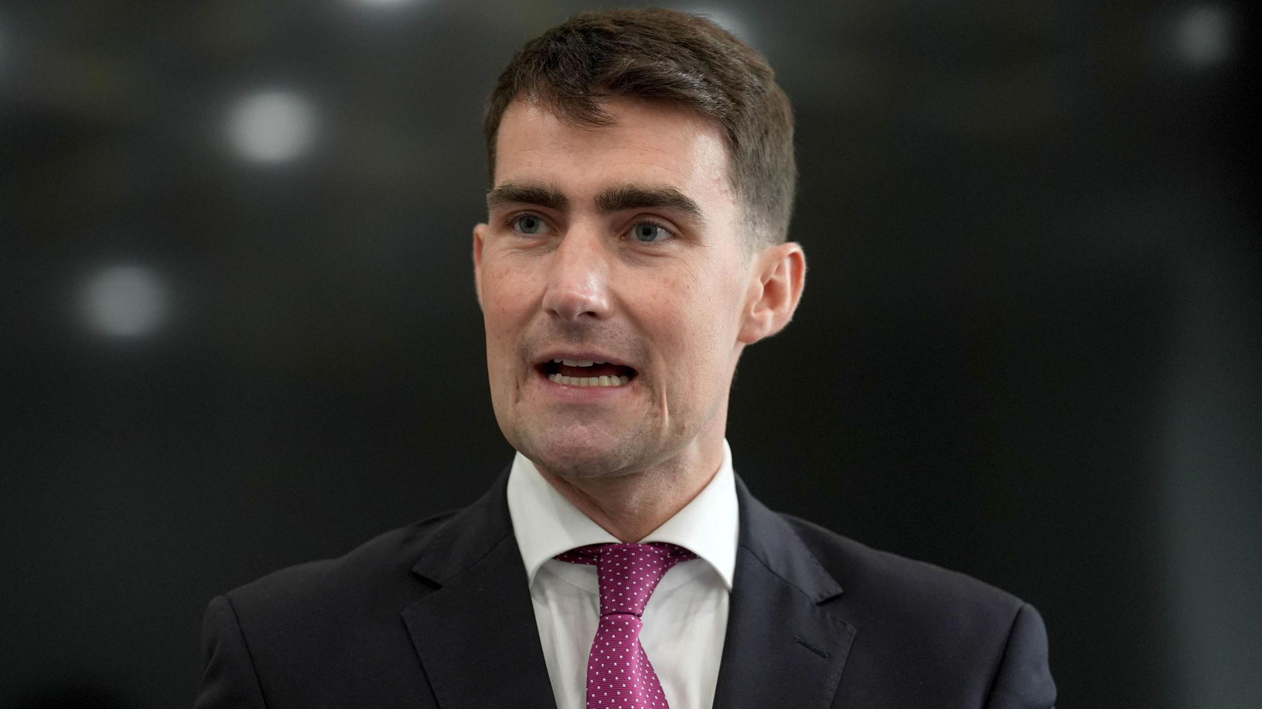 Jack Chambers in black suit, white shirt and pink tie. He has black hair.