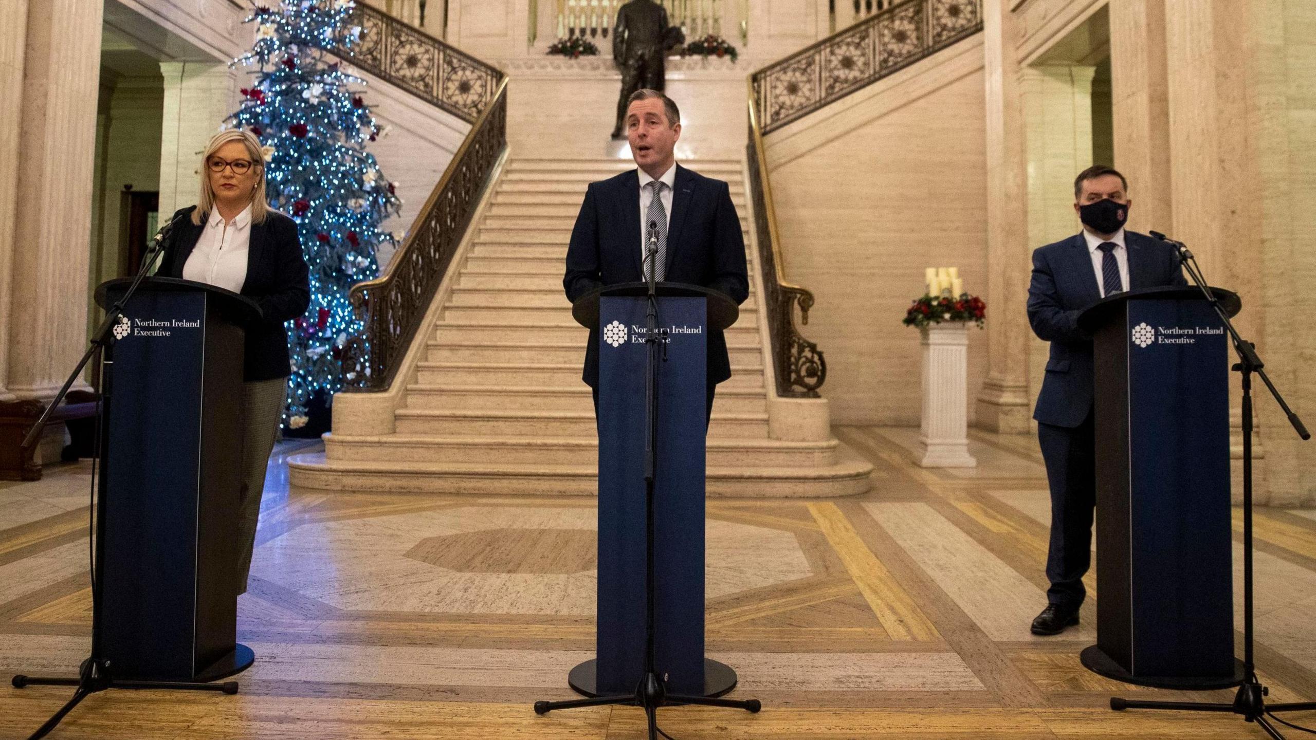 Michelle O'Neill, Paul Givan and Robin Swann standing behind lecterns in Stormont. A Christmas tree is behind them.
