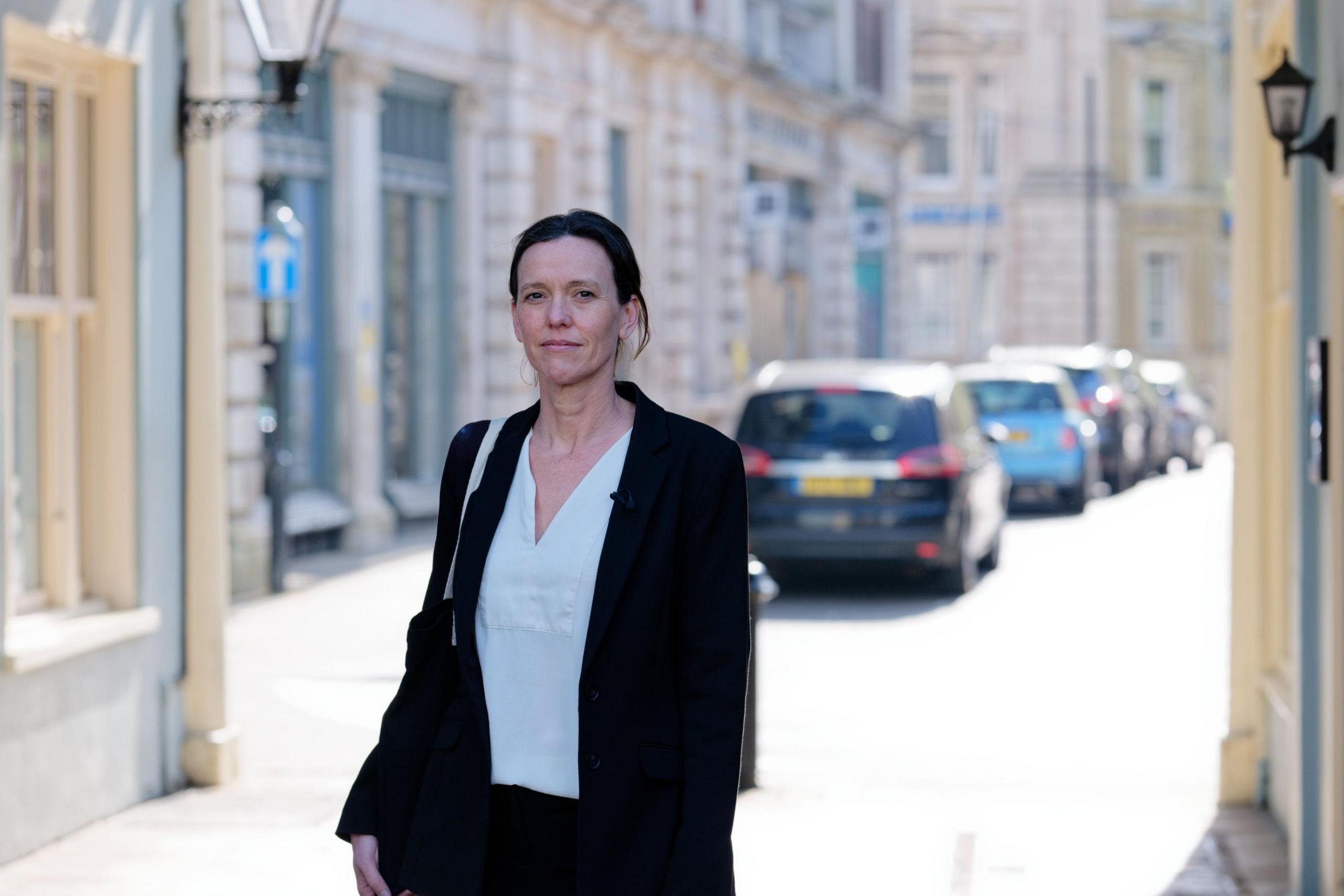 Lucy Davies walking towards court. She has dark brown hair tied back in a ponytail. She is smartly dressed with a white blouse and a dark suit jacket with a bag over her right shoulder.