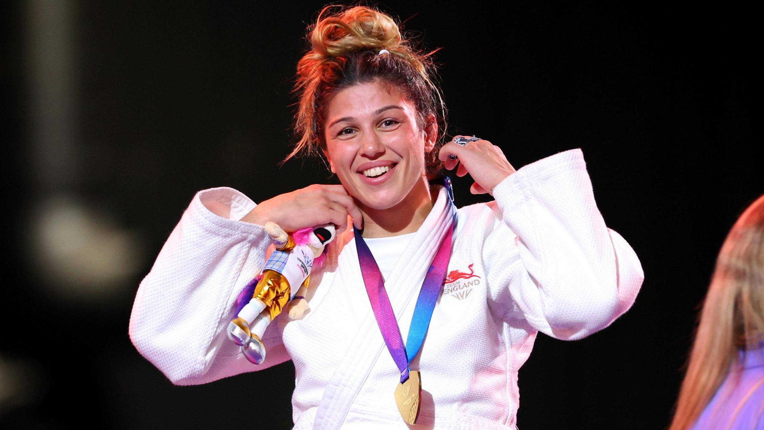 Acelya Toprak, wearing England's white judo robe, smiles with her gold medal around her neck on the podium