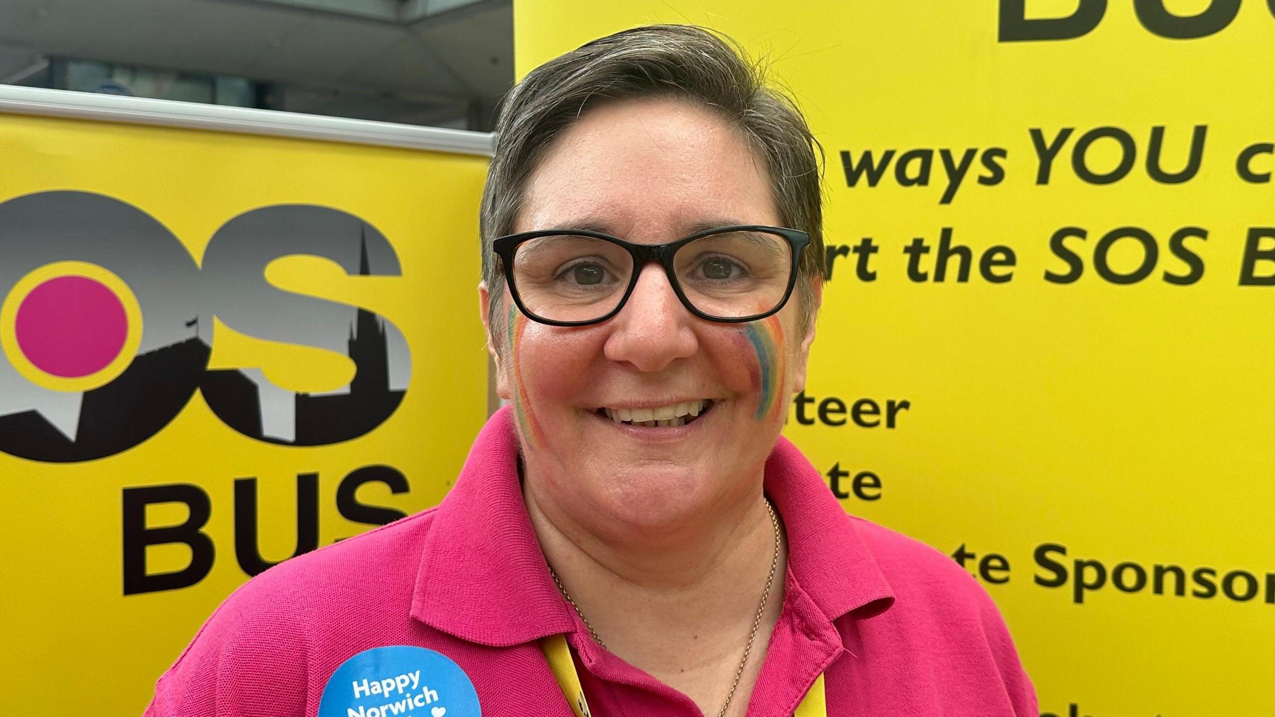Tracey Slater is standing in the middle of the image wearing a pink top. She has a yellow lanyard around her top and is standing in front of a yellow banner. 