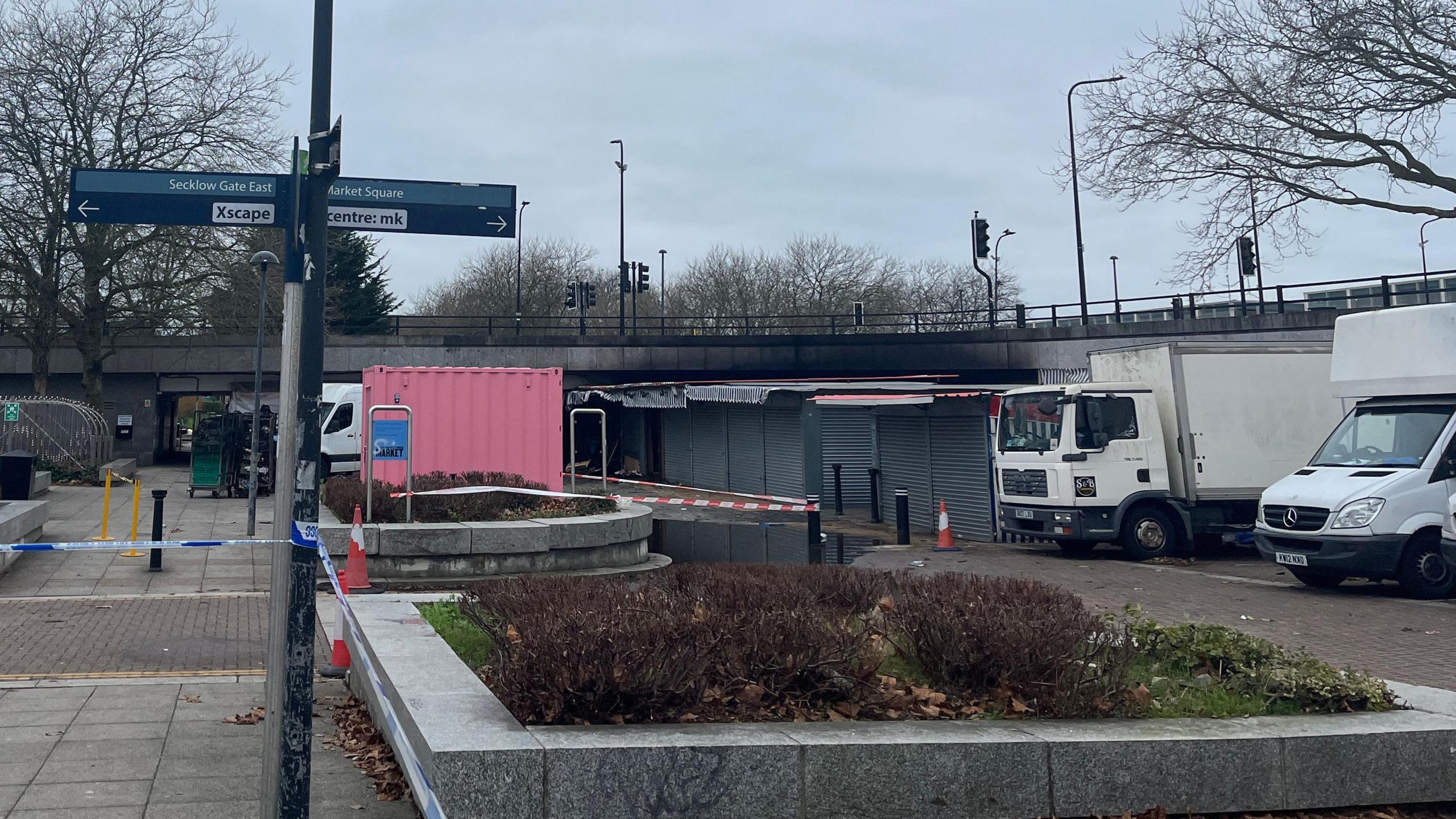 Milton Keynes Market which is a range of covered stalls. A sign can be seen on the left hand side of the picture pointing to the left which says Secklow Gate East and Xscape. The sign points right to Market Square and "centre: mk".  The bridge/raised road above the market can be seen with black scorch marks on its side. Red and white tape can be seen around one section of the market stalls and a blue and white police cordon tape is also in place. 