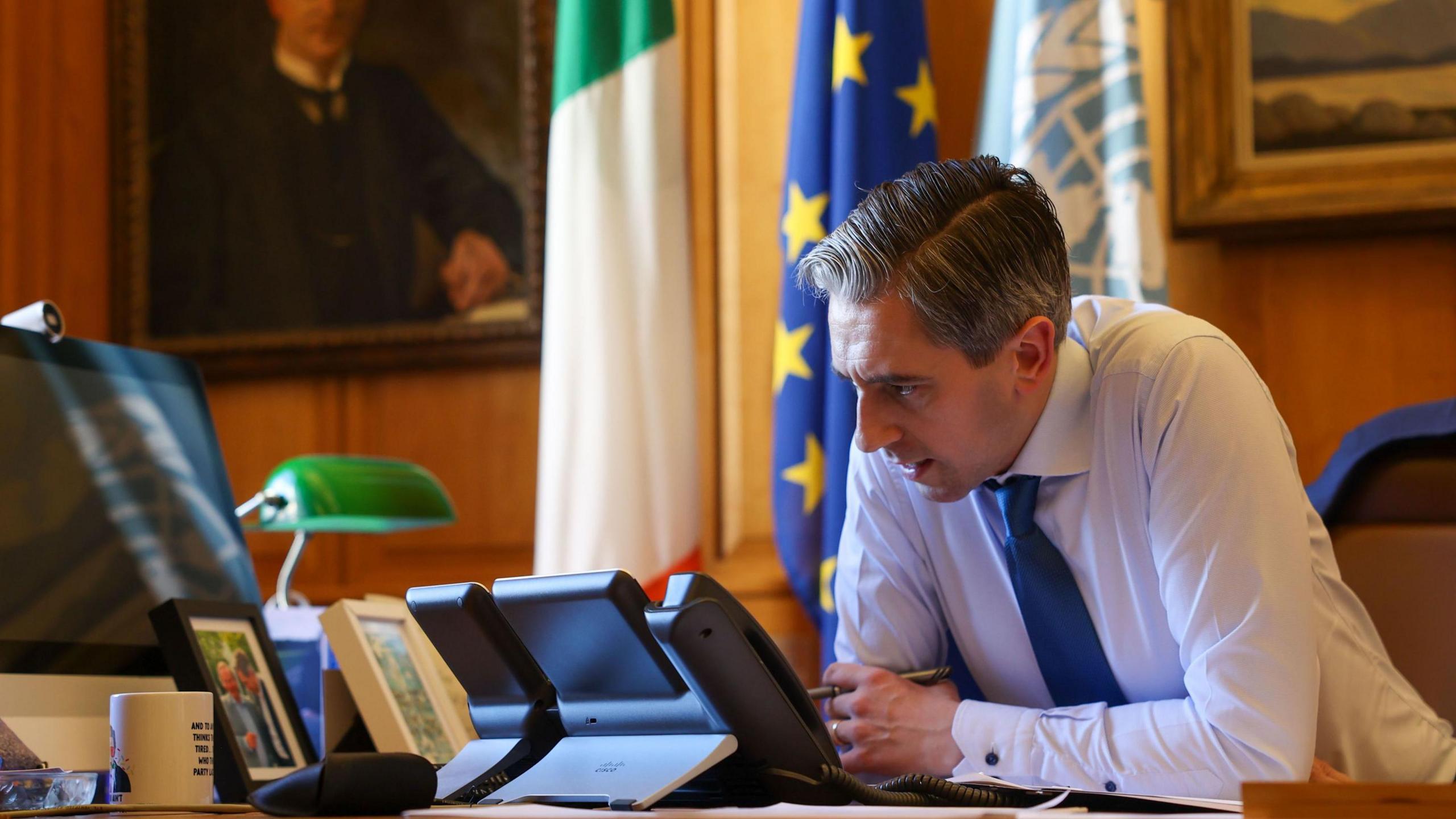 The Taoiseach sitting at a desk wearing a white shirt and blue tie. He is speaking into a black phone. On the desk is a computer screen, framed photographs and a white cup. In the background there are three flags hanging down the wall and two large portraits.