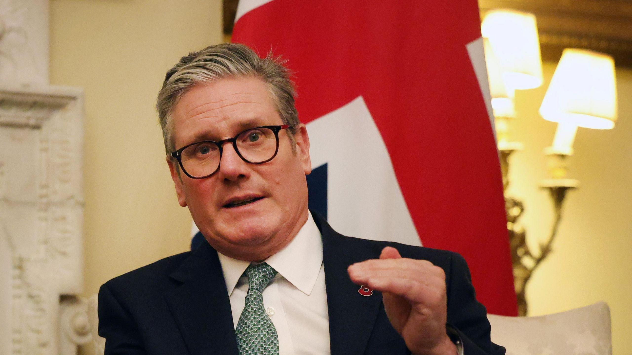 Sir Keir Starmer speaking while seated n Downing Street. He has one hand raised while speaking and the Union Jack flag behind them.