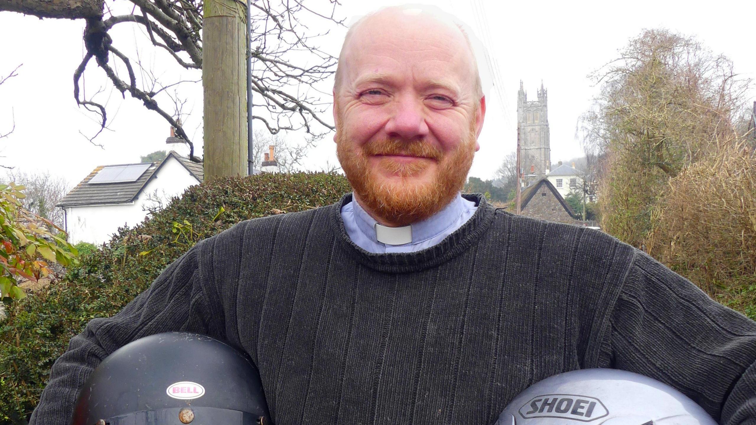 A man wearing a dog collar and a jumper smiles at the camera as he holds two motorbike helmets