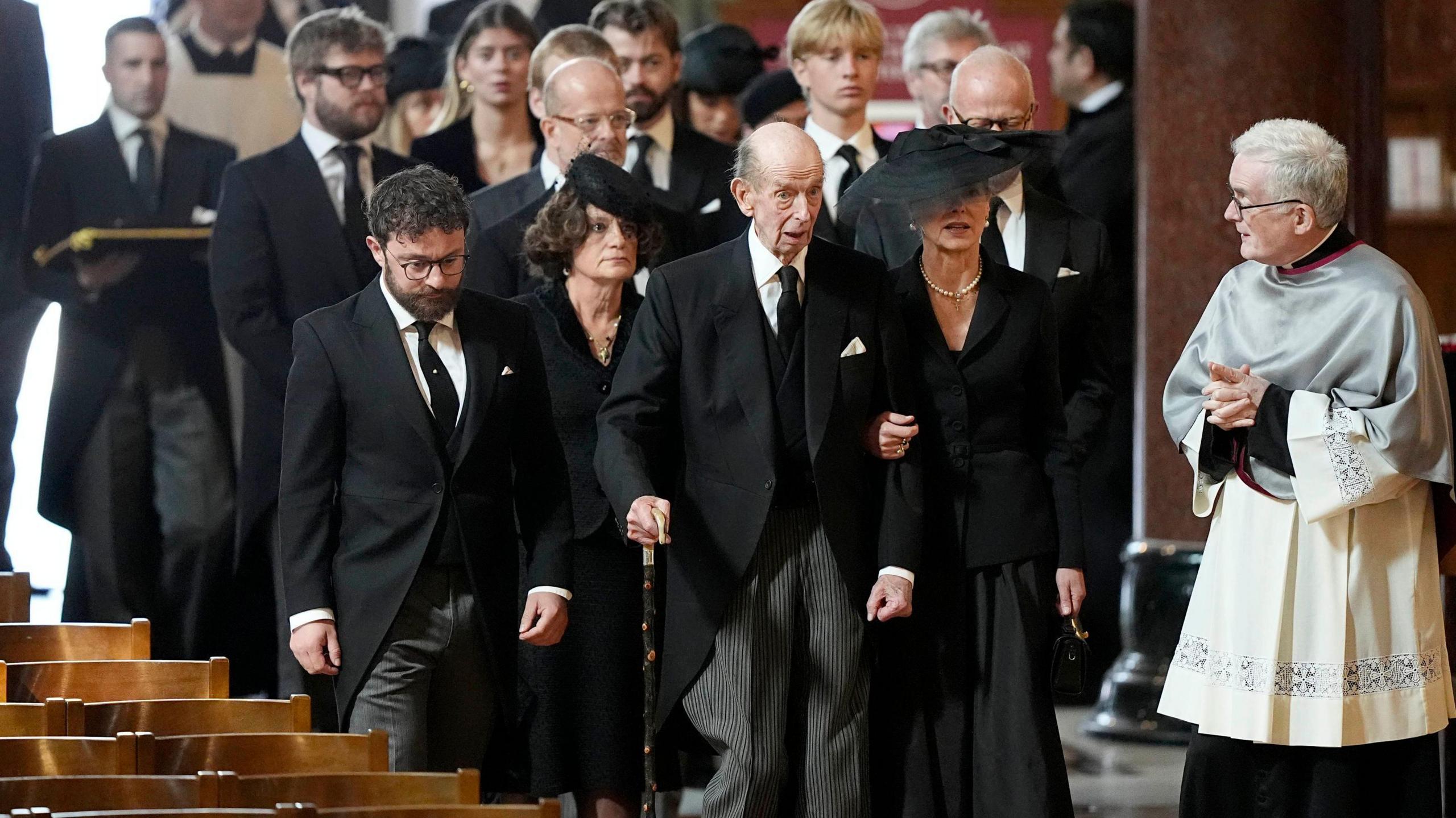 The Duke of Kent at the funeral of his wife Katharine. He walks aided by a stick and is wearing a black morning suit. He is accompanied by his children and grandchildren.