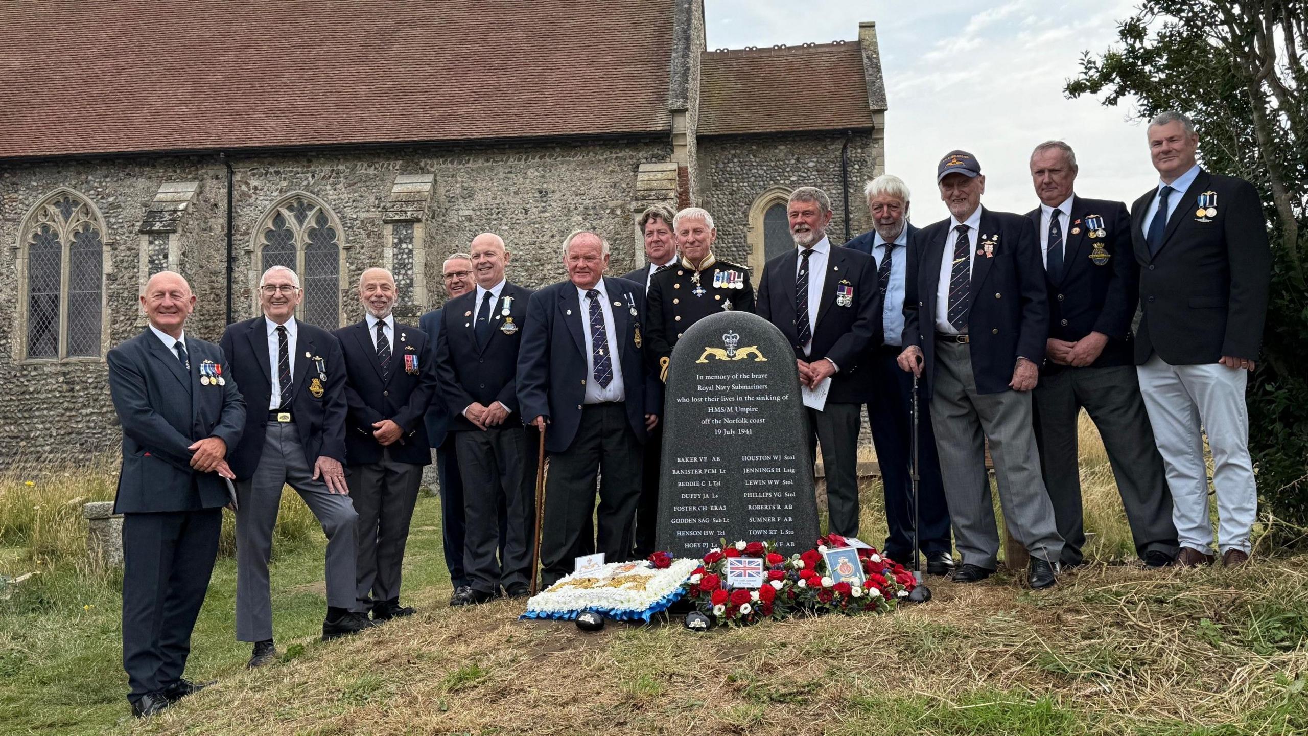 Men dressed smartly in navy blazers, with medals,  line up behind a new gravestone-shaped memorial, with Mundesley church behind them 