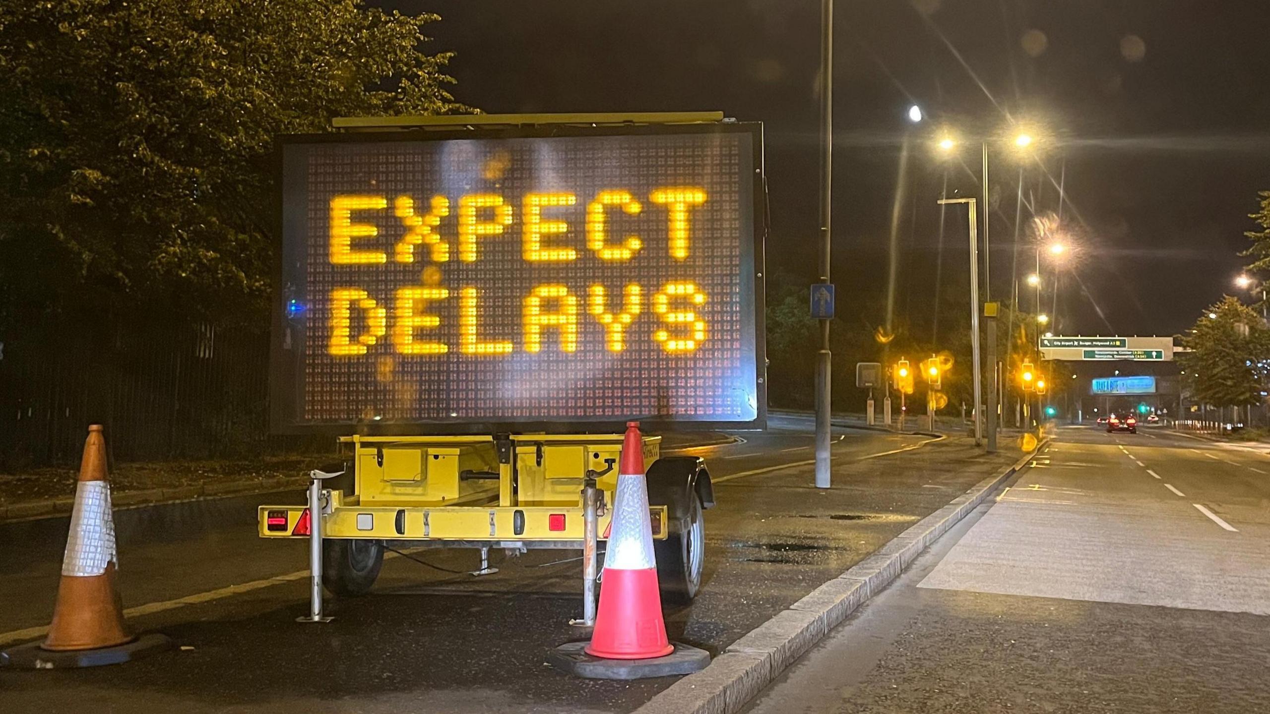 A black sign saying "expect delays" is lit up. It is on the side of the road and there are two traffic cones in front of it.