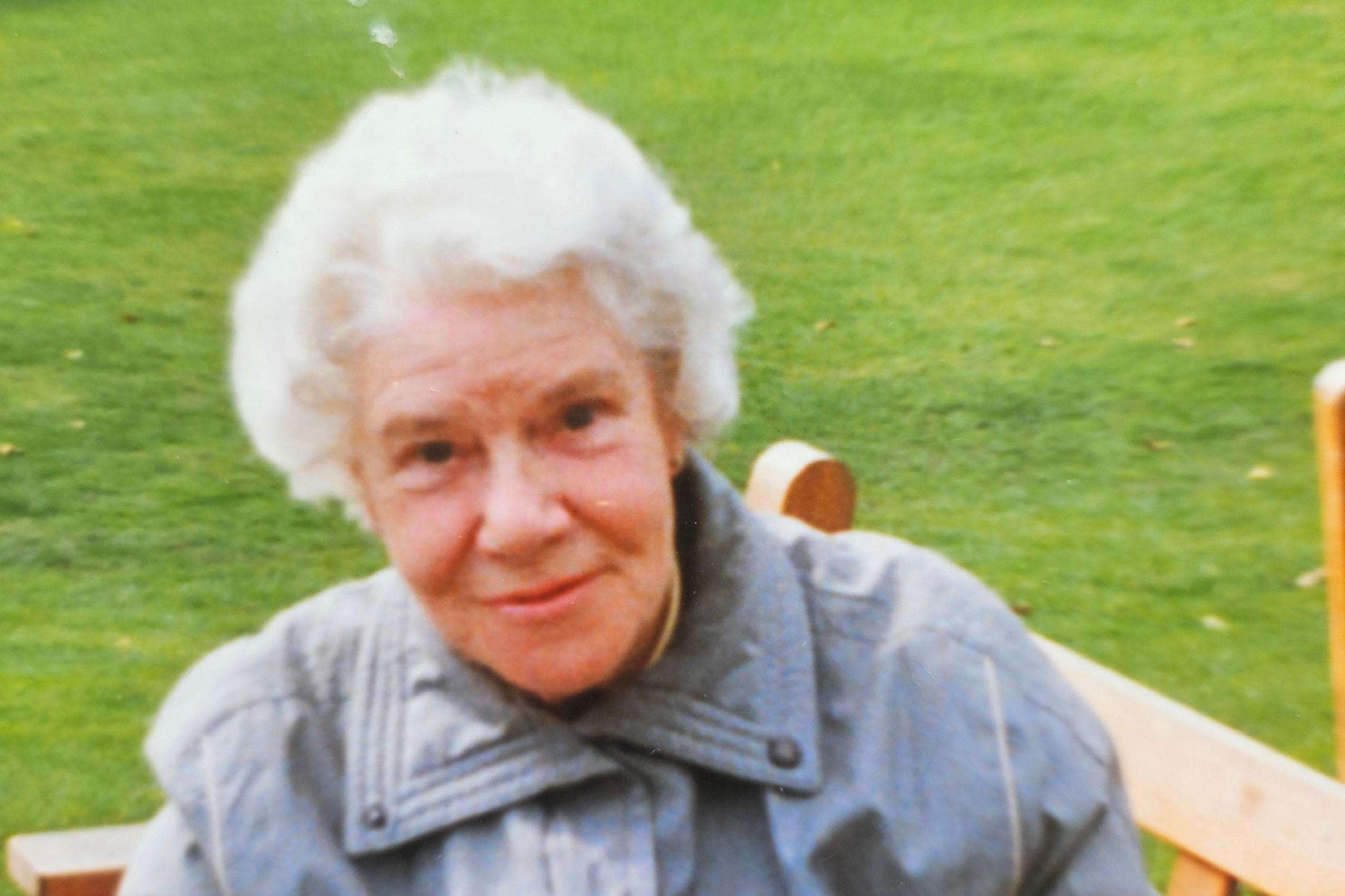 A photo of an elderly lady with short white hair wearing a grey coat, smiling slightly at the camera. She sits on a bench with grass behind her.