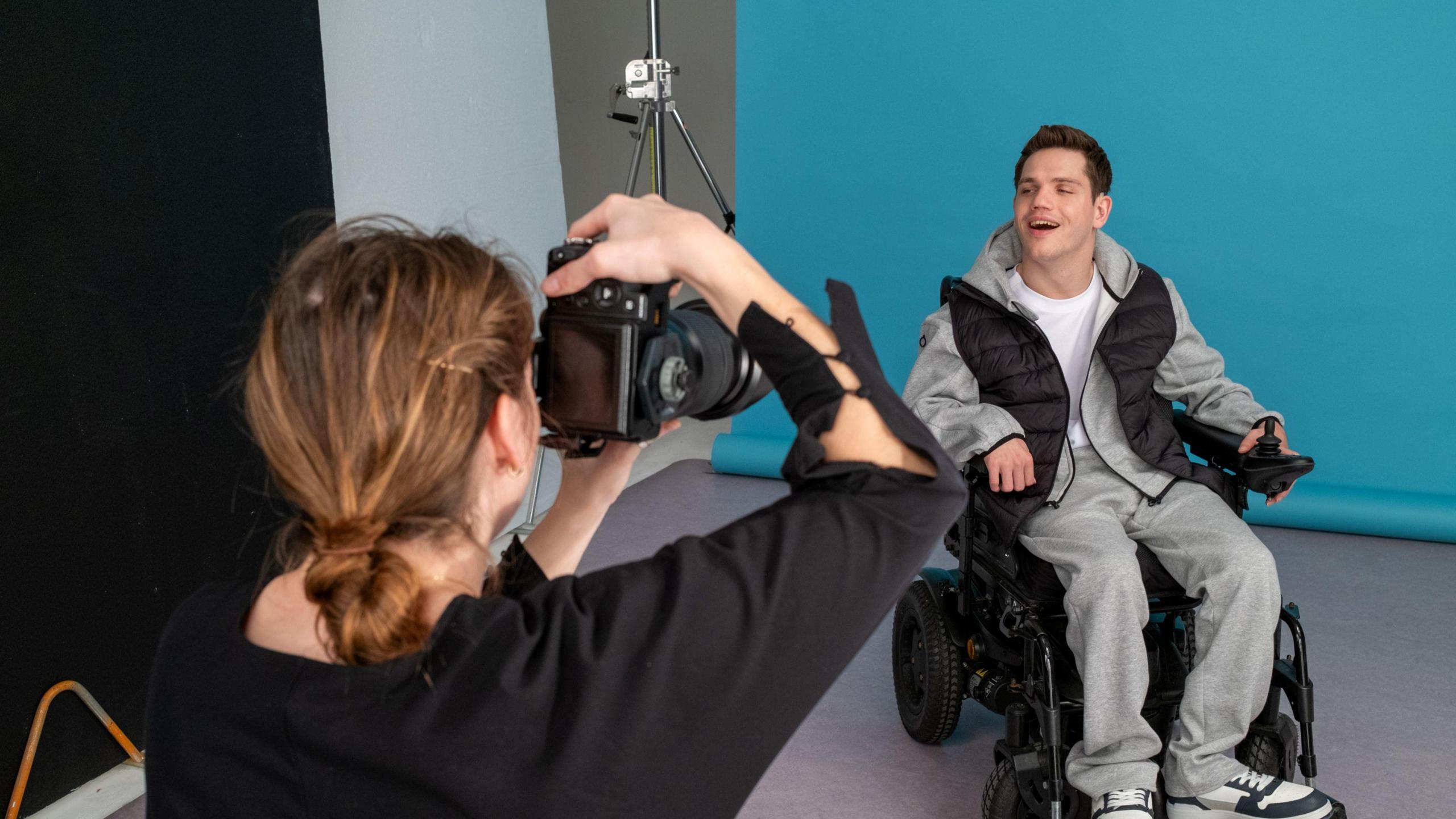 A man, Elliot Caswell, is sitting in an electric wheelchair in front of a blue backdrop in a photography studio. He is wearing a grey tracksuit and a black gilet and is smiling at the camera. A woman in a black top is taking his photo with a large camera.