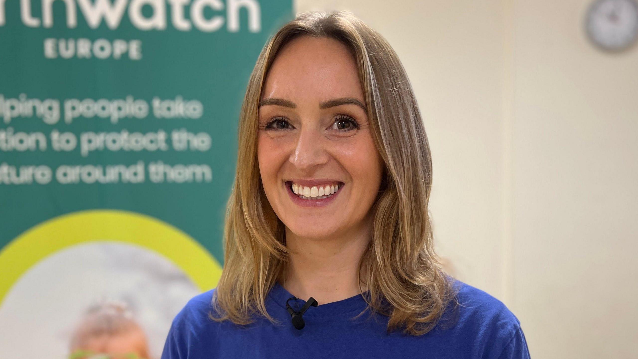 Dr Sasha Woods is in the middle smiling at the camera. She has shoulder length light brown hair and brown eyes, and is wearing a blue T-shirt. She is stood next to a sign on the left. 