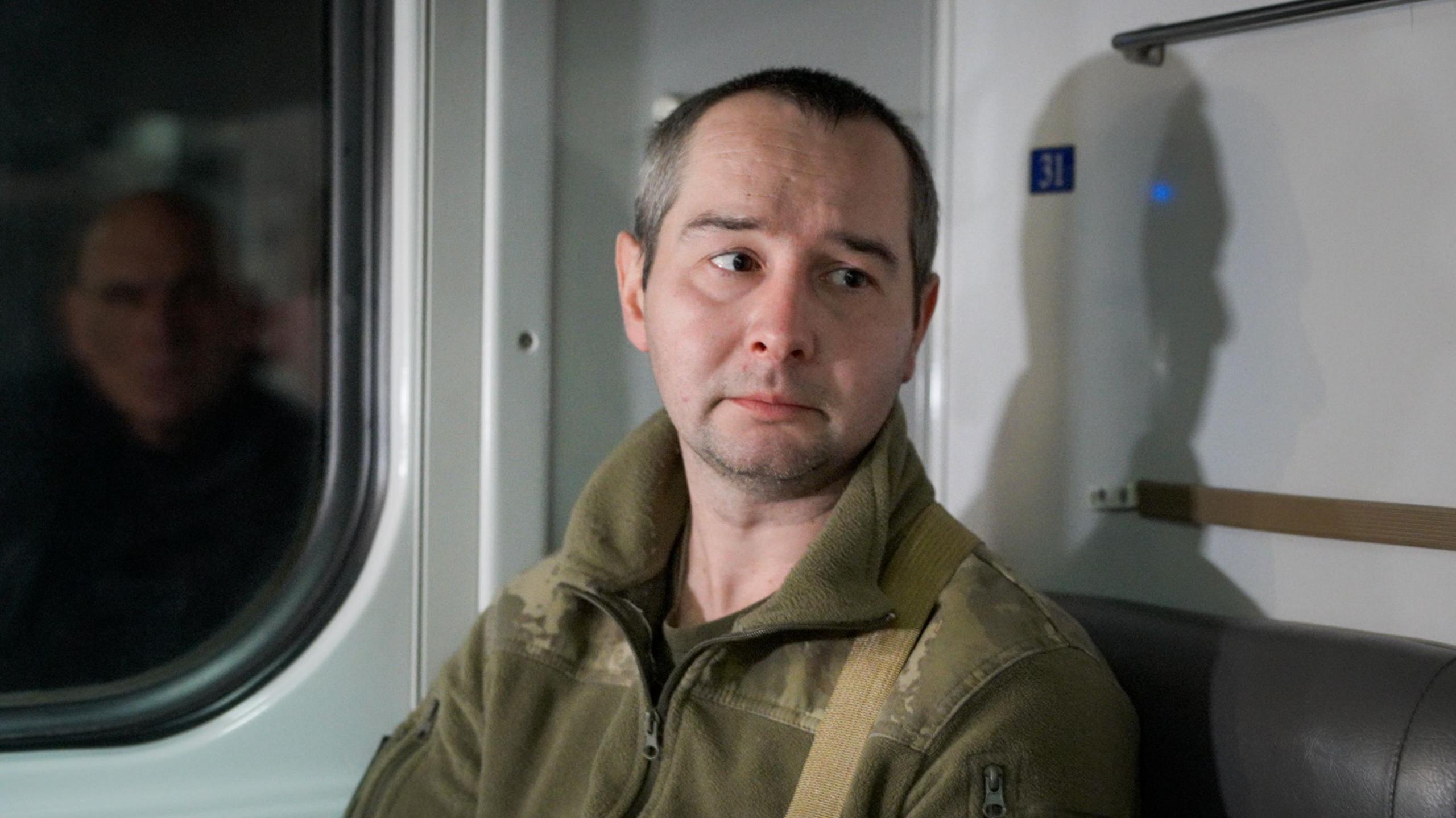 Denys, sitting on a train, looks down away from the camera, lips downturned. He is wearing military fatigues. 
