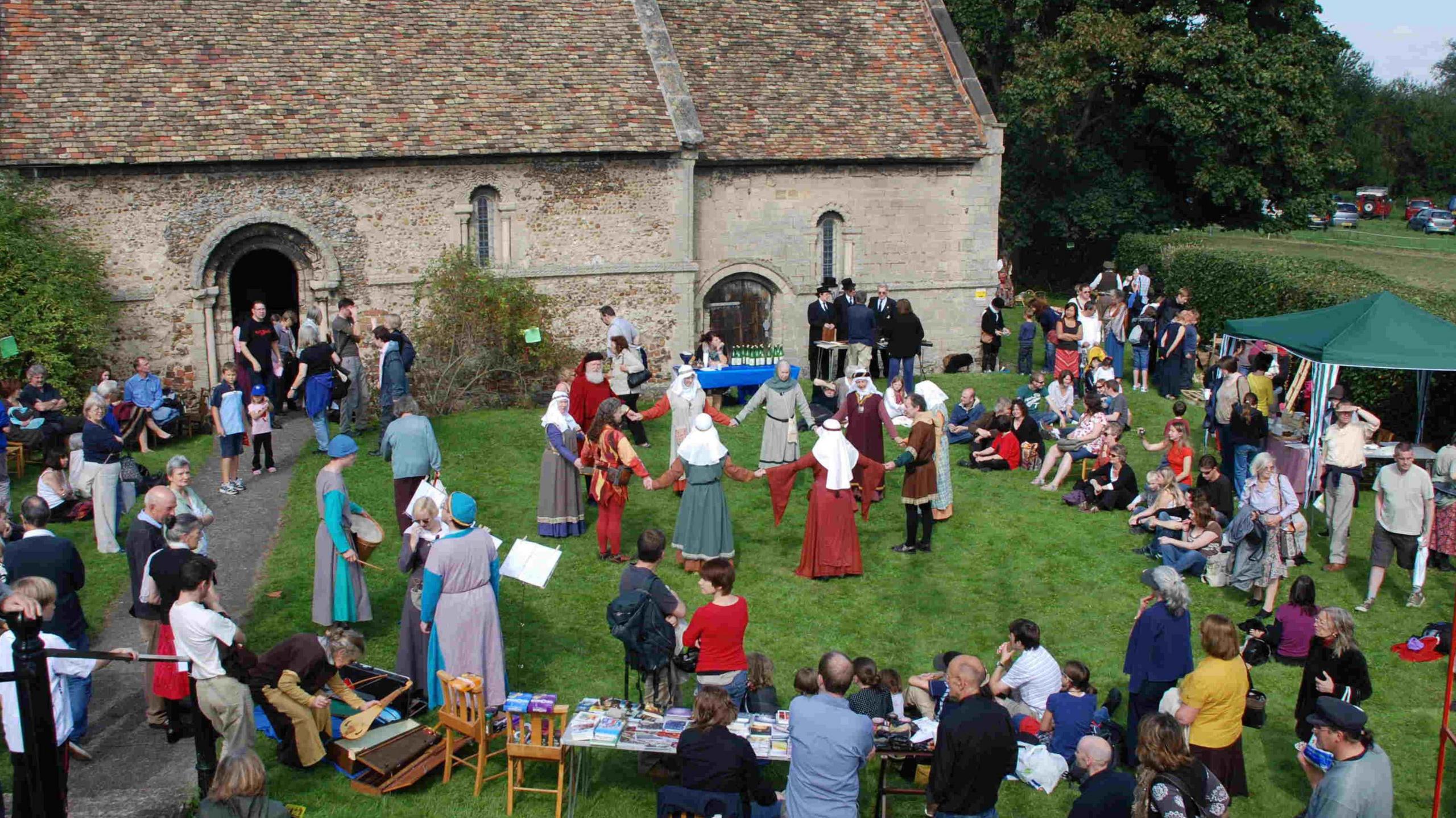 Aerial view of a previous fair outside the  Leper Chapel, a rectangular building made from pale stone with narrow windows and a slated roof. On the grass are people stood in a circle holding hands, they appear to be starting a dance. Behind them are some musicians who are also in medieval dress, with long garments over the backs of their heads and long dresses. Surrounding them is another circle of people - visitors to the fair who are watching the performance.
