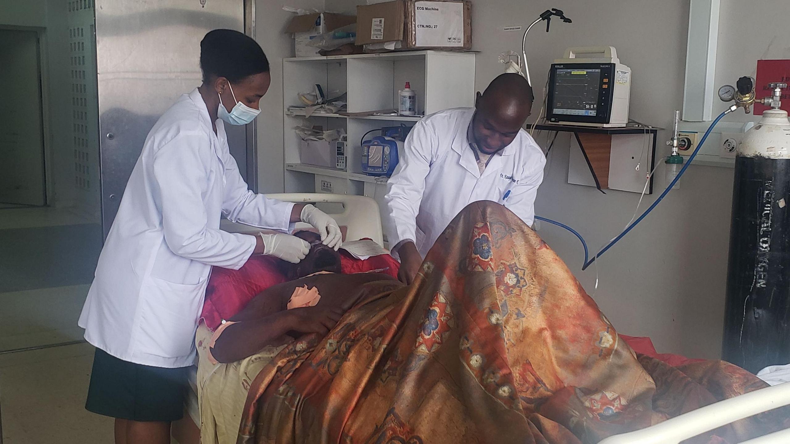 Lawrence Mugejera lying in a hospital bed, with two health workers in white overcoats attending to him