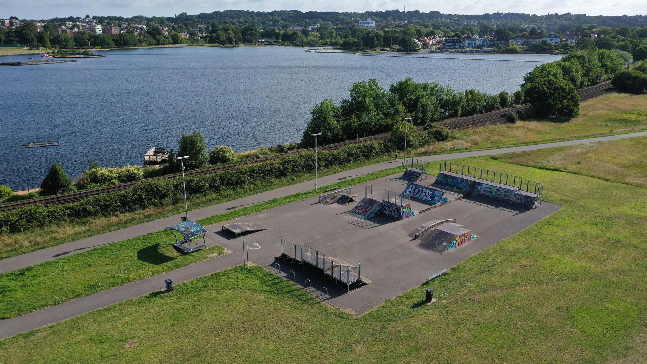 Green space in park next to a lake with a skate park with ramps set in a space of rectangular tarmac.