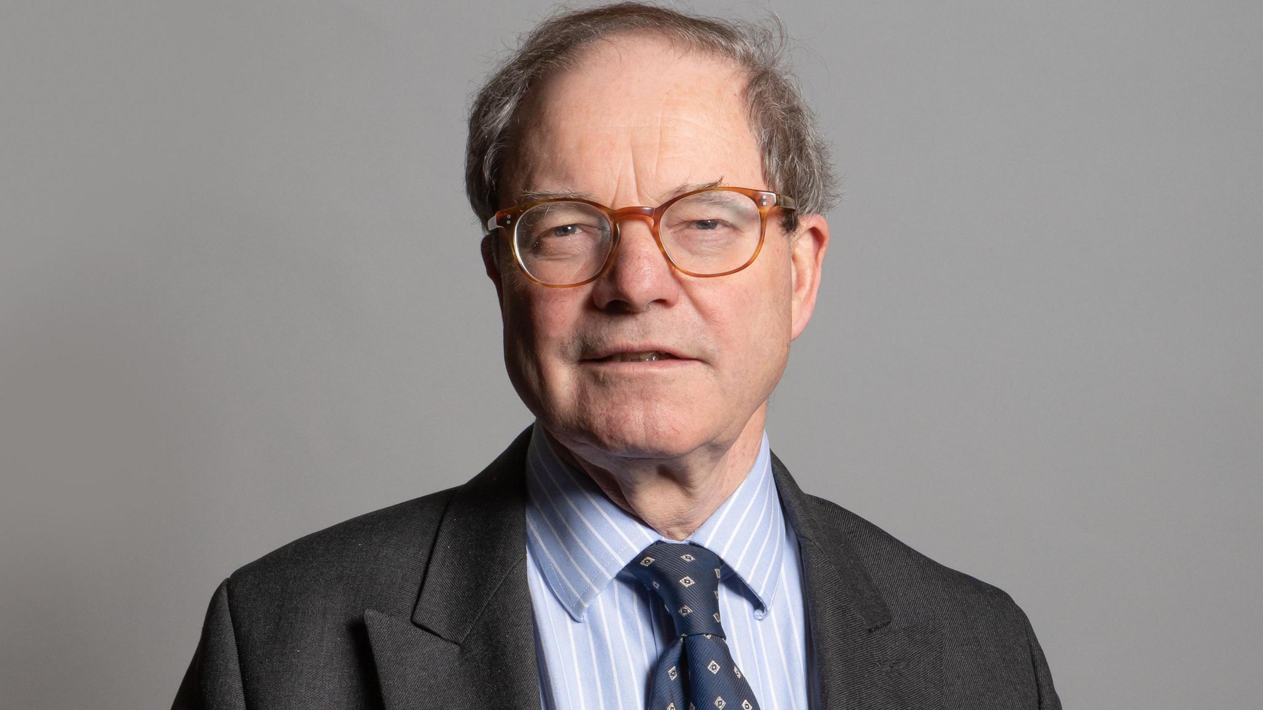 Sir Geoffrey Clifton-Brown. He has short grey hair, glasses and is wearing a suit. He is looking at the camera and smiling slightly. 