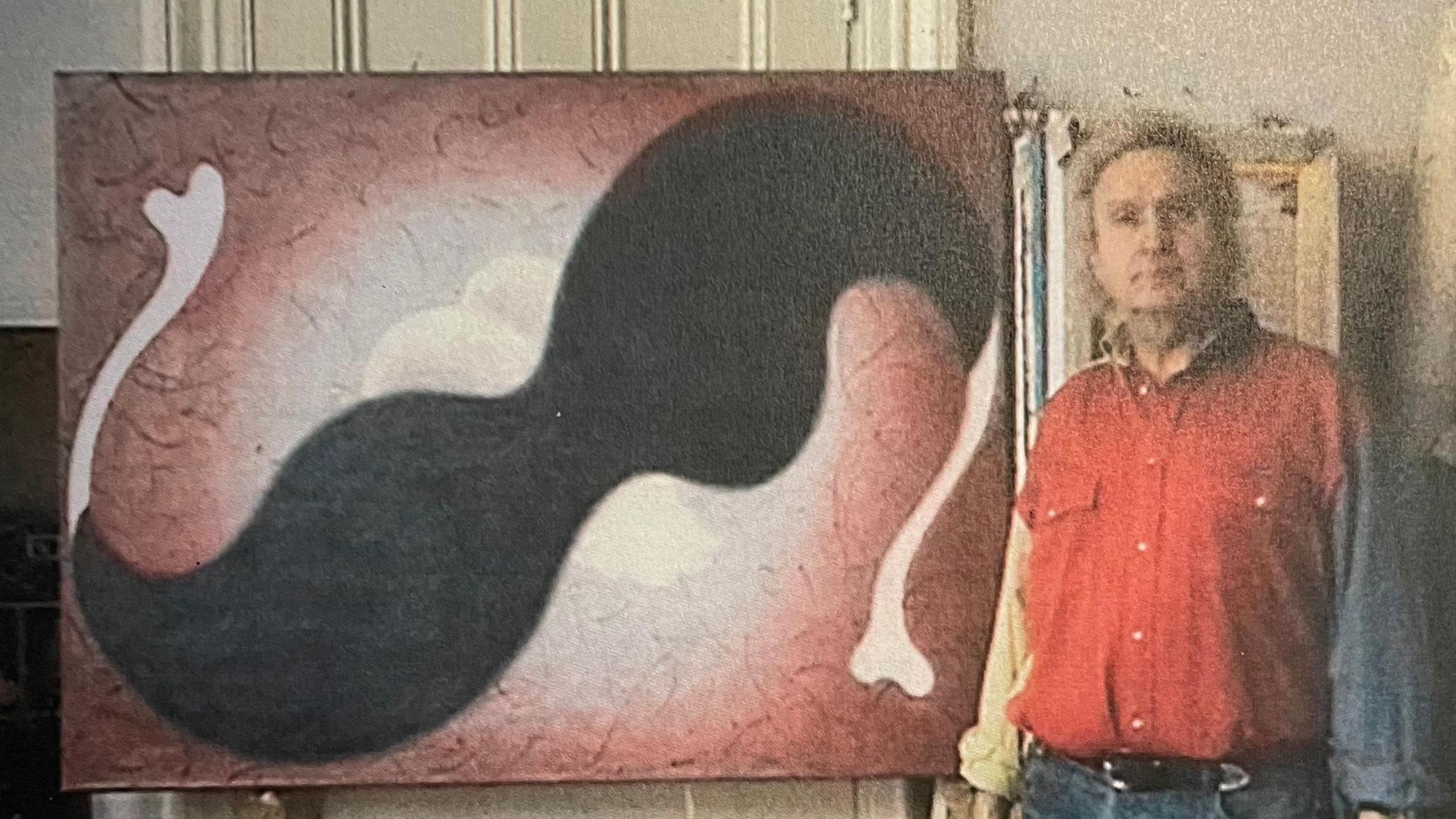 A grainy image of Henry Orlik when younger in a red shirt standing next to one of his surreal artworks, depicting a warped pair of sunglasses.