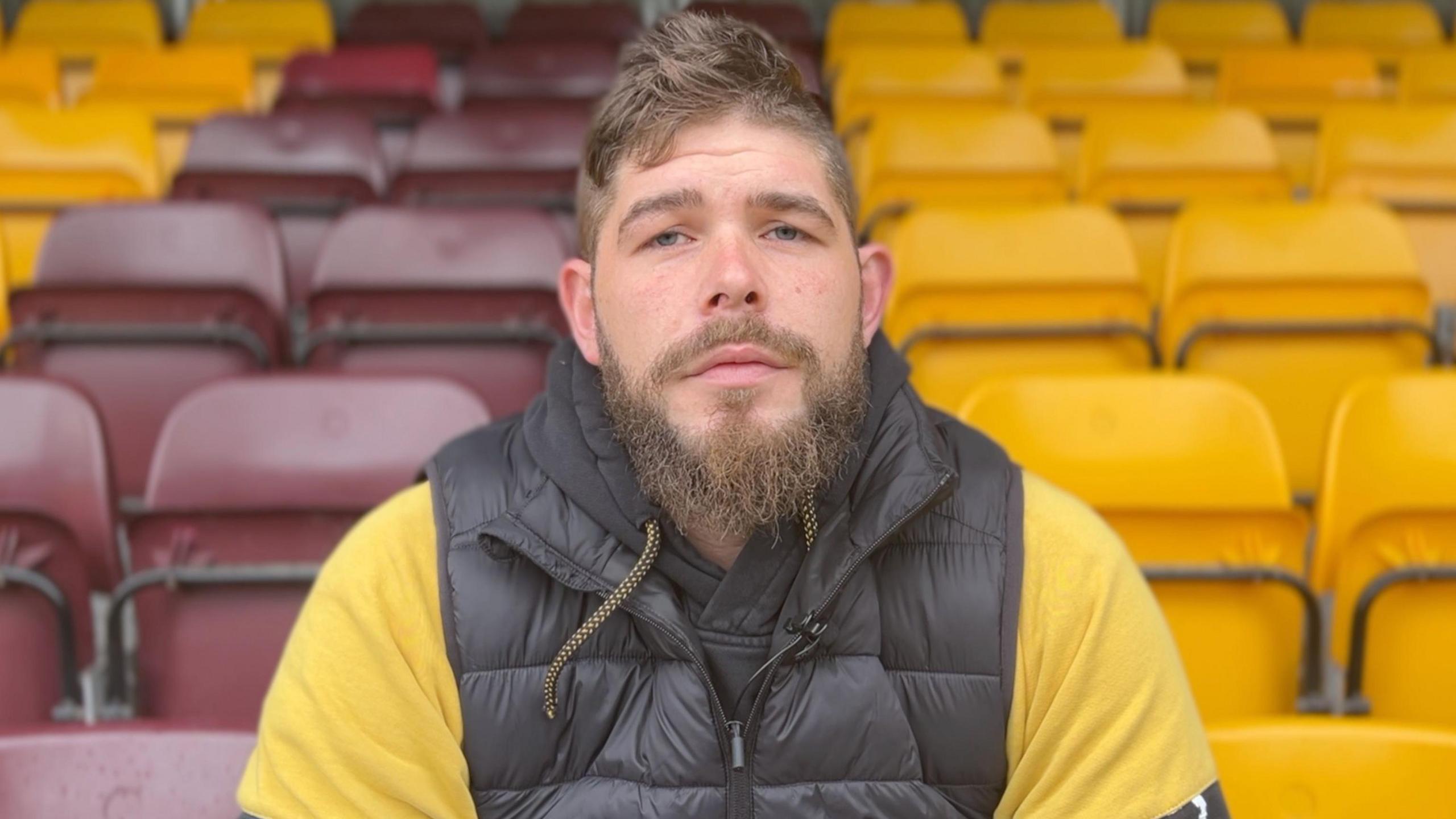 A man with brown hair and a beard sits with empty rugby stadium seats behind him. He is wearing a black sleeveless jacket and yellow top underneath.