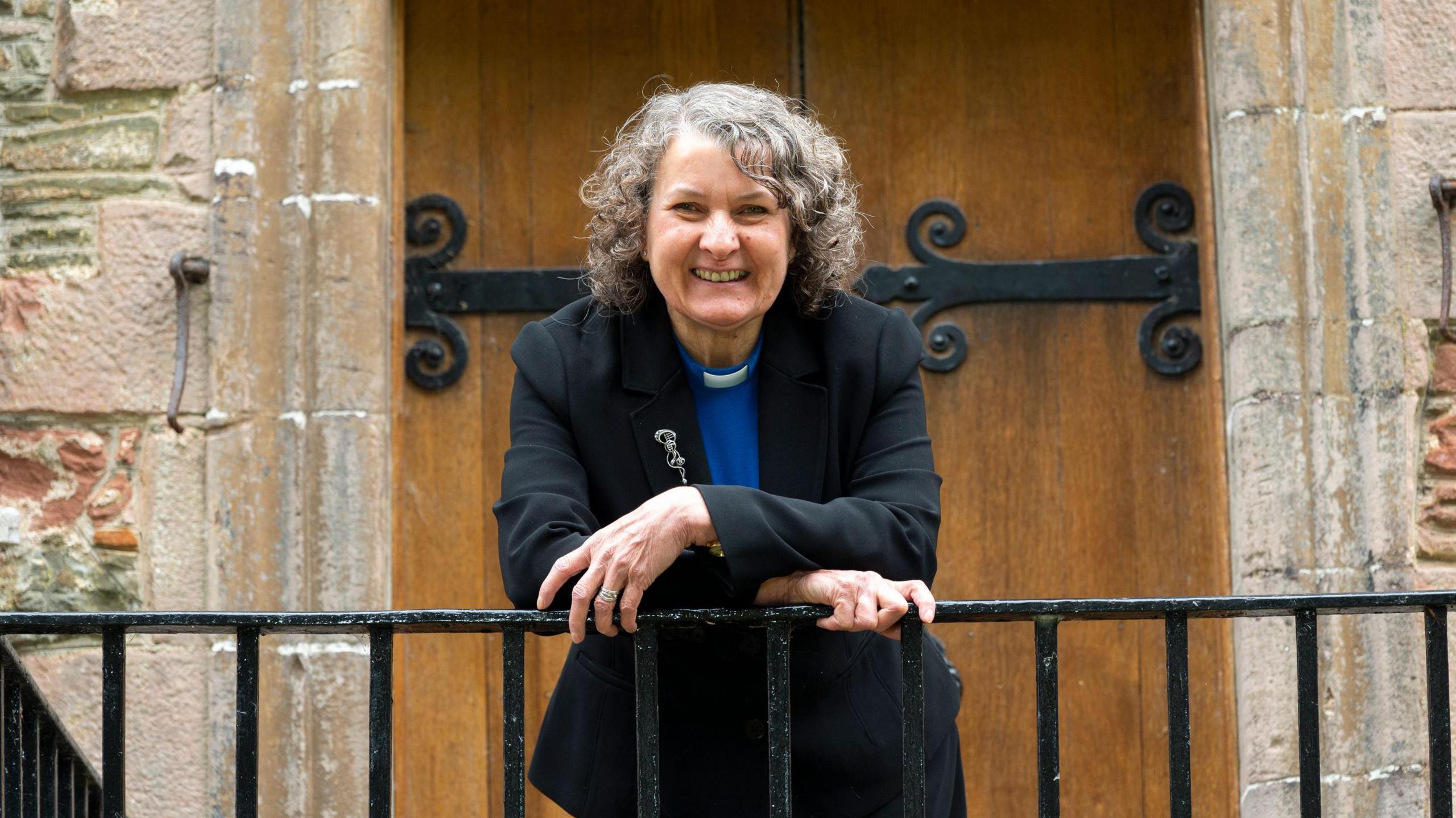 A female minister with grey hair leans over a metal railing smiling in front of a large wooden door.