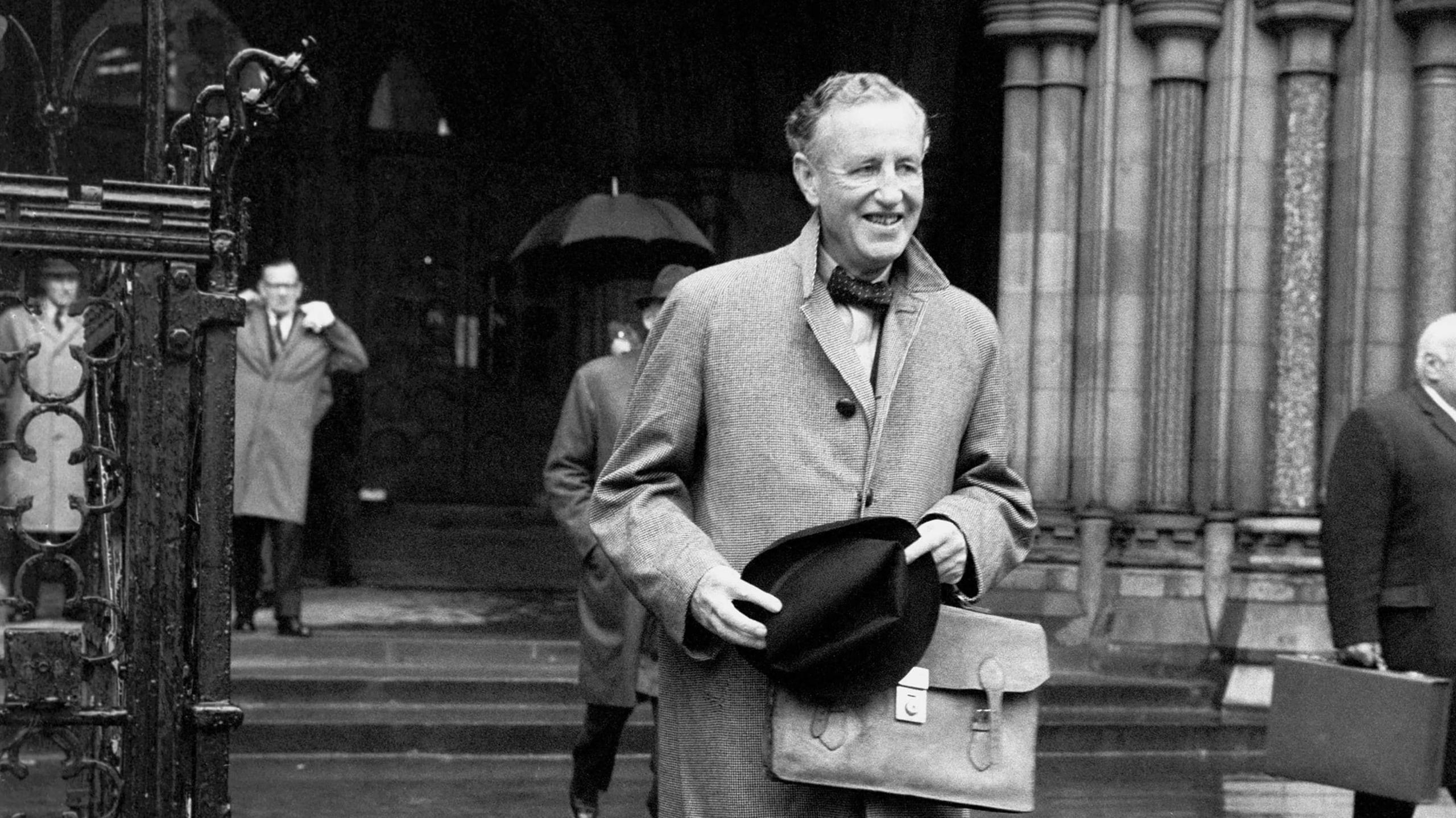 Black and White image of Ian Fleming outside a big building wearing a coat and holding his hat in 1964
