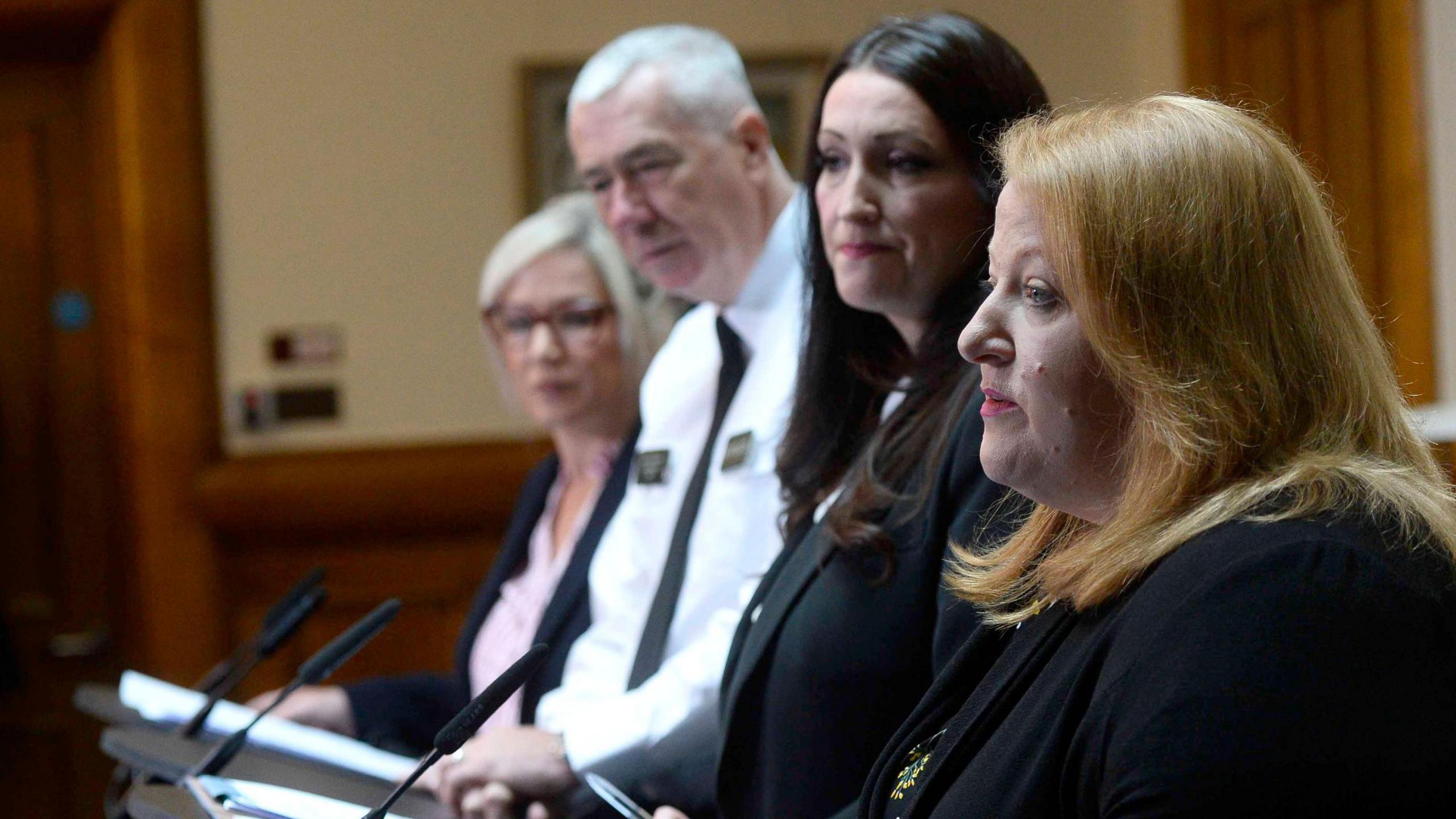 Naomi Long speaking at a podium with Emma Little Pengally, Jon Boucher and Michelle O'Neill beside her looking concerned 