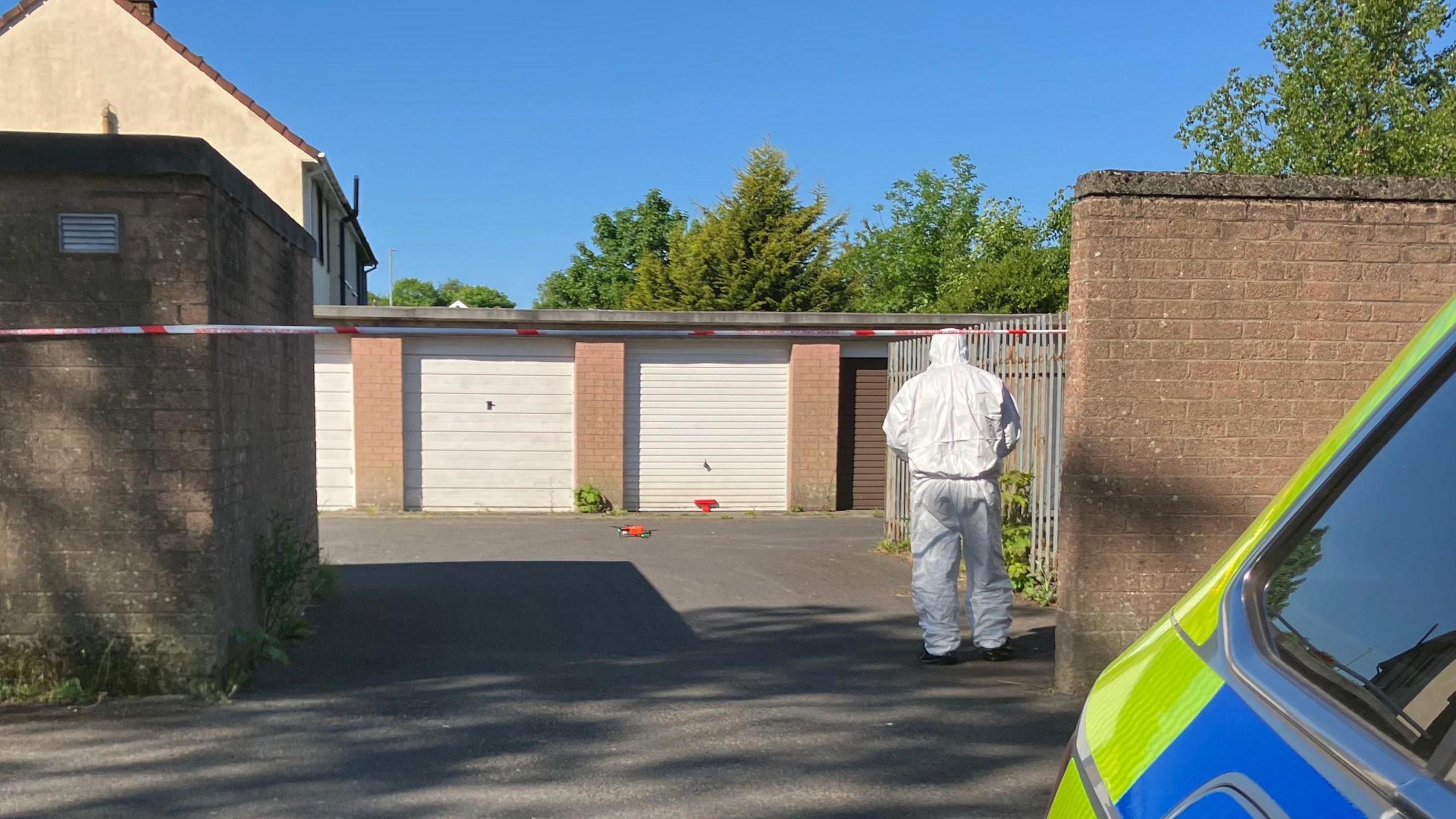 A forensic officer in a white suits walks towards a row of garages with his back to the camera. There is a white and red police tape blocking of entry to the garages which have white and brown roller doors. There is a brown brick wall next to the forensic officer. Beside the camera is a yellow and blue police car.