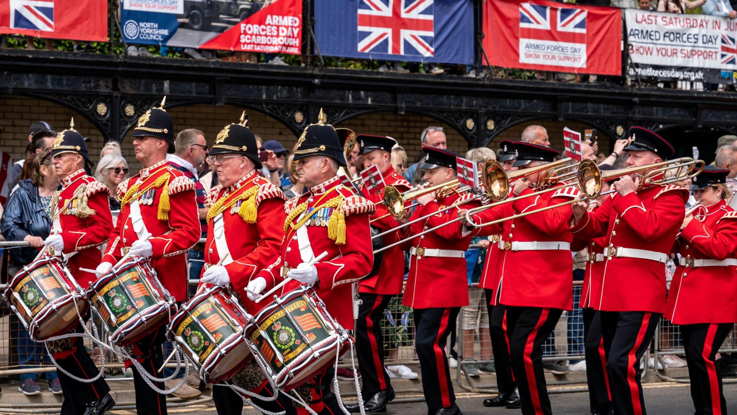 A parade of a military band with drums and men in red uniforms marching past a large crowd