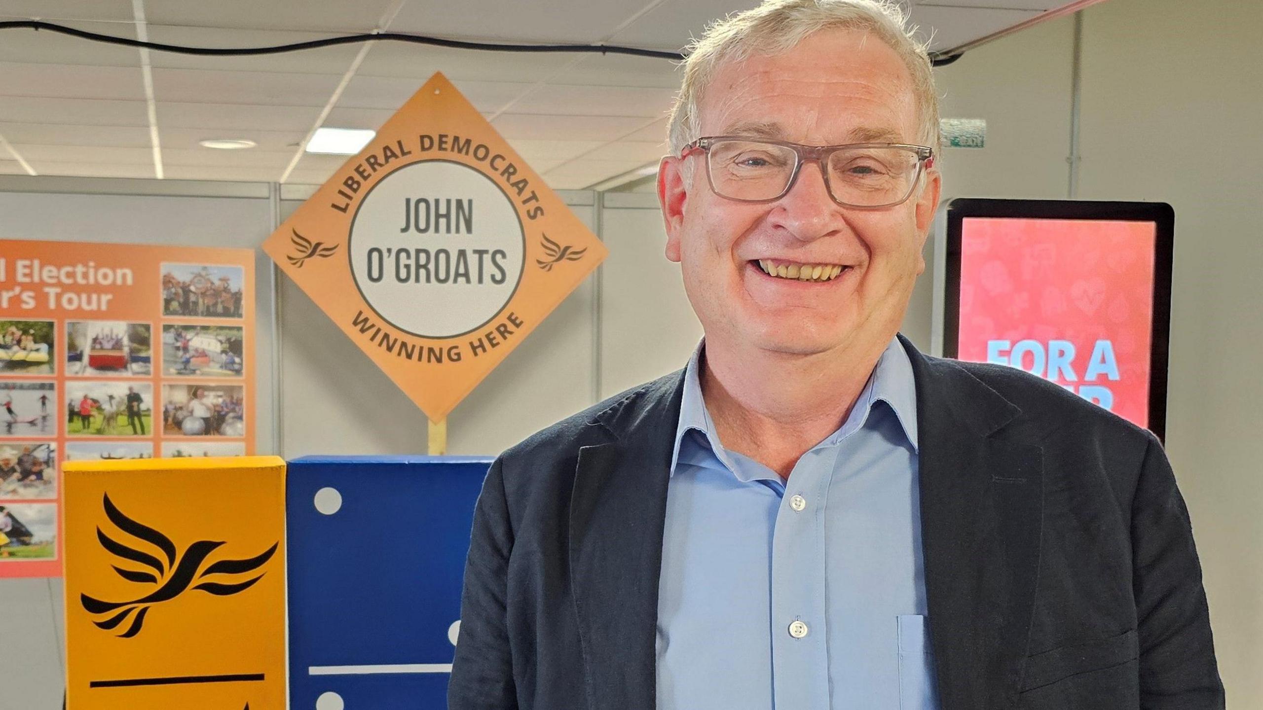 Graham Colley smiles at the camera in a blue open-collared shirt and a dark jacket. He has grey hair and wears glasses. He is standing in front of various Lib Dem banners and posters at the conference.