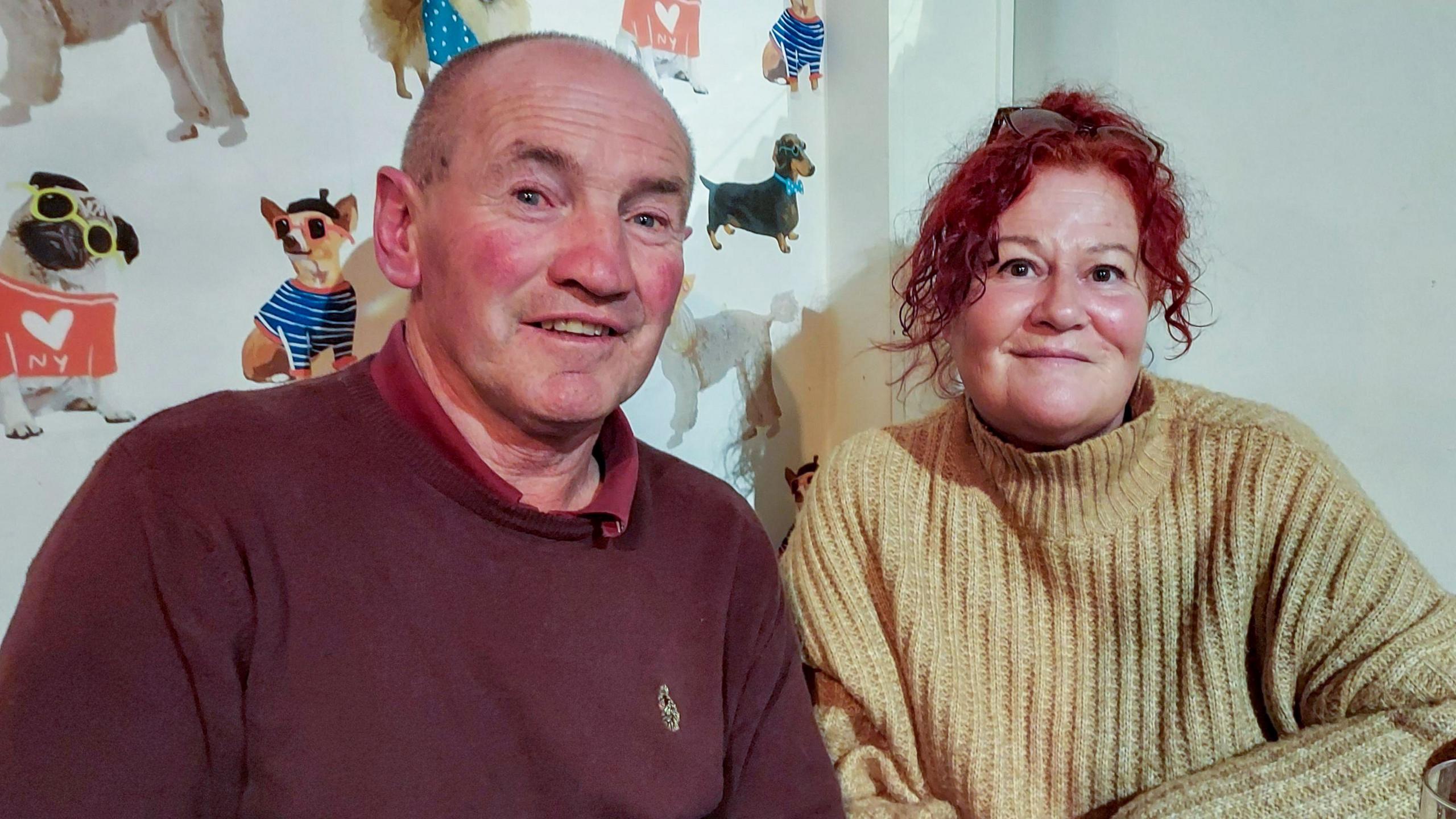 John Kelly with shaved hair wearing a brown polo top and jumper and Carole Kelly with auburn curly hair and black glasses resting on her head wearing a beige roll neck jumper. They are sitting in a pub and both are smiling.