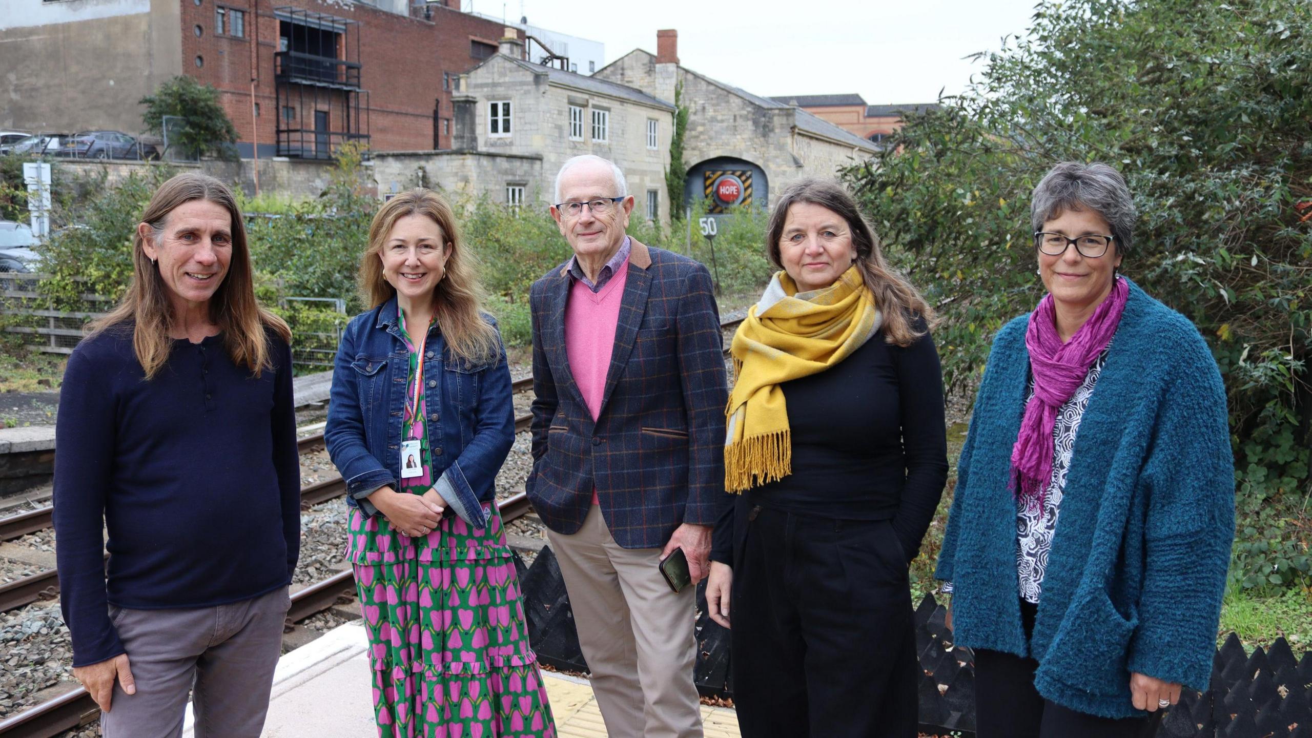 A group of five people, two men and three women, standing on the rail station platform outside the Brunel Goods Shed. All are smiling at the camera.