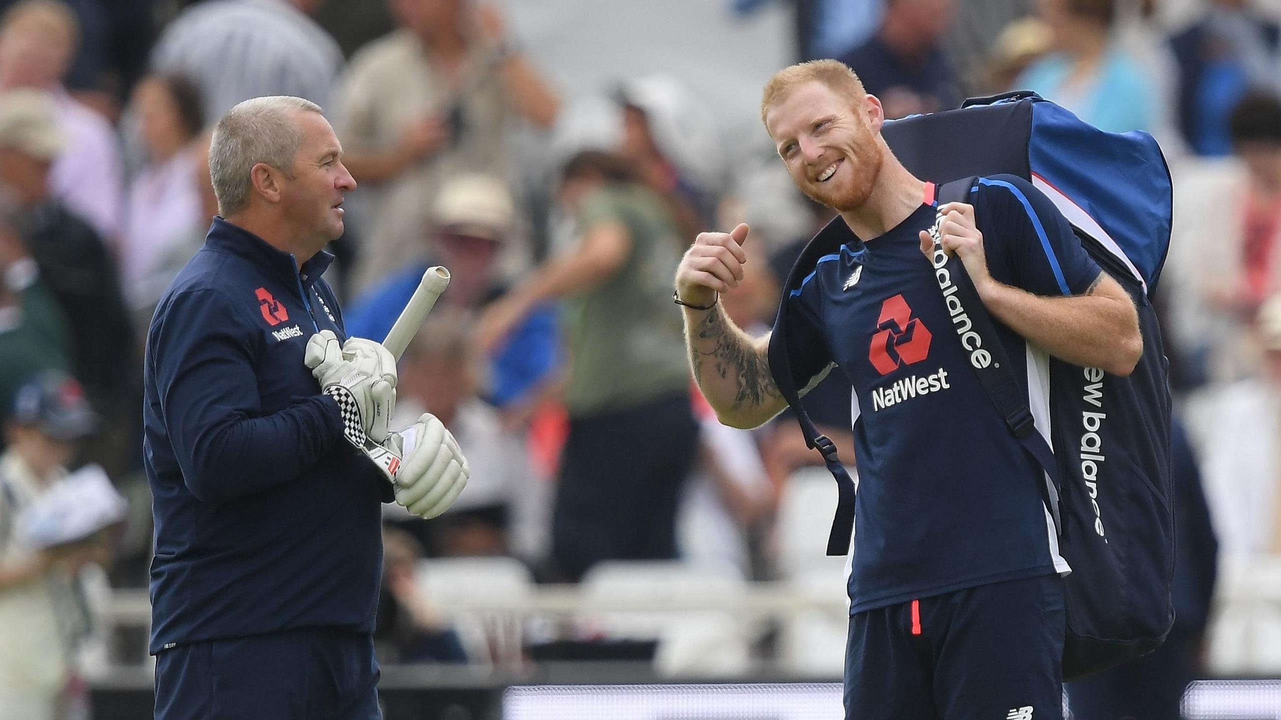 Paul Farbrace, wearing batting gloves and a bat under his left arm, shares a conversation with a laughing Ben Stokes, who has his big cricket bag across both shoulders 