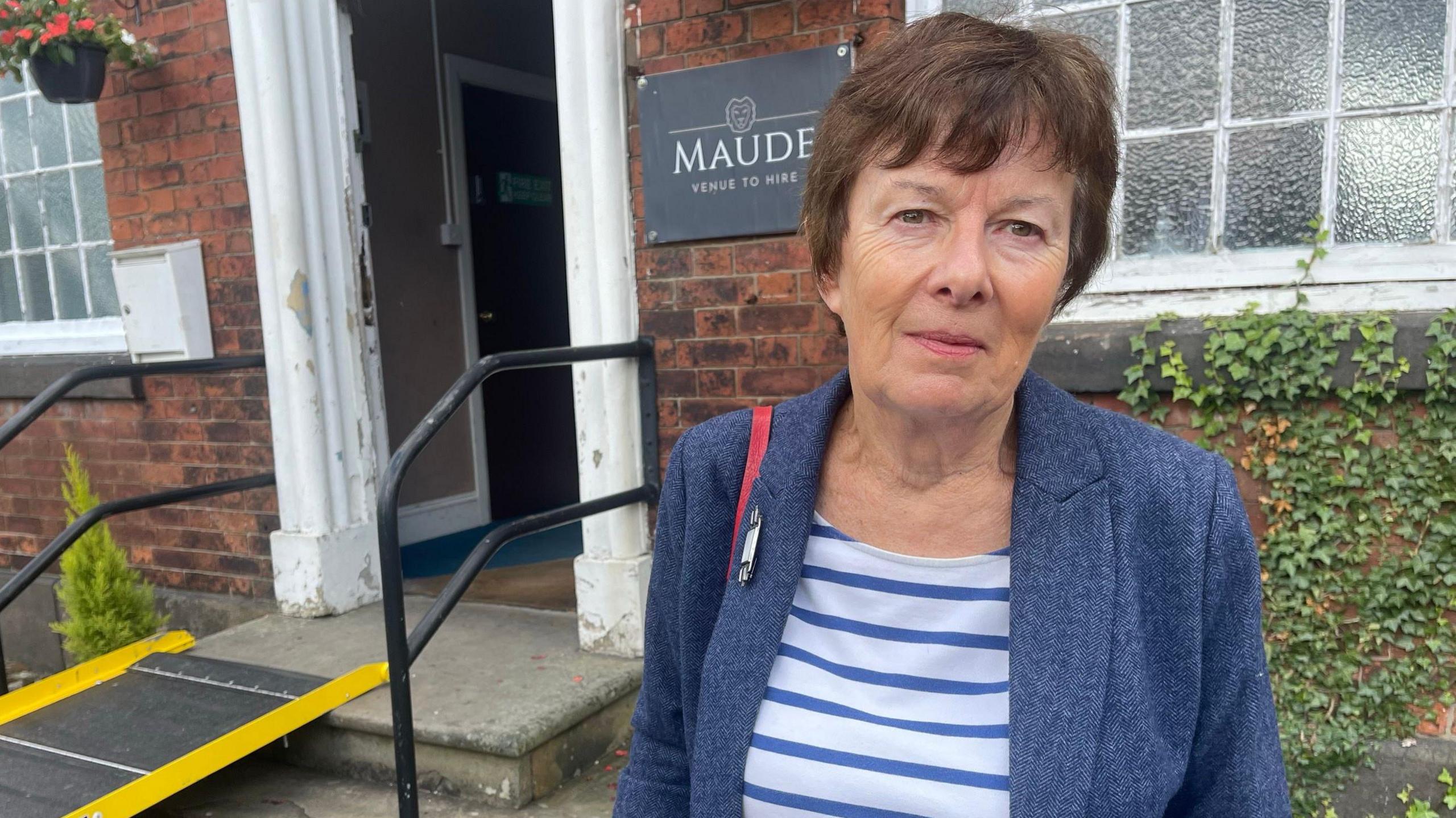 Charlotte Atkins wears a blue jacket over a blue and white striped top. She has short dark hair and the red strap of a bag is visible over her right shoulder. She stands in front of the entrance to a building which has a ramp going up to the front step.