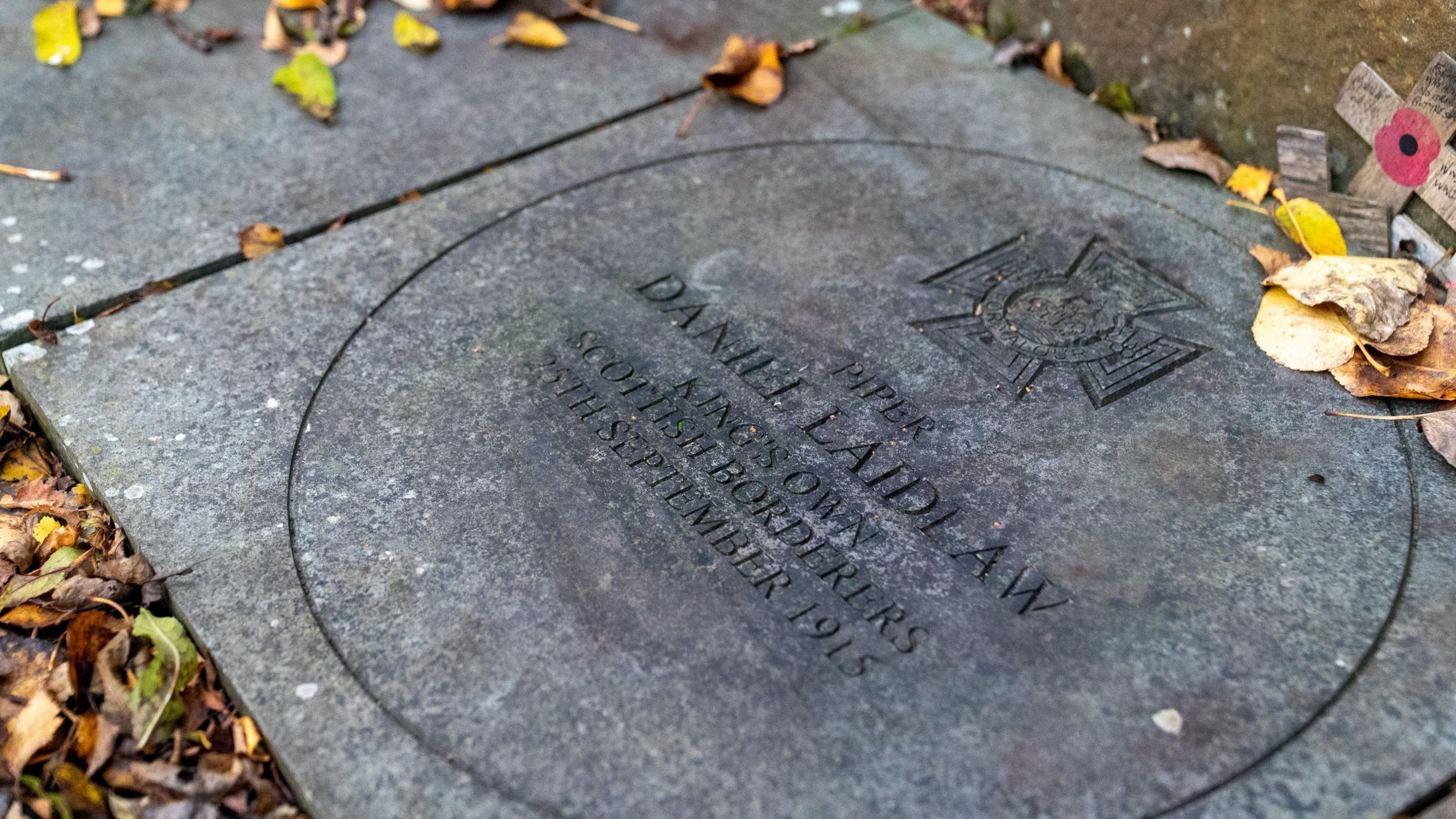A circular stone plaque engraved with the name Piper Daniel Laidlaw and details of his service lies among fallen autumn leaves. A small wooden remembrance cross with a red poppy rests nearby.