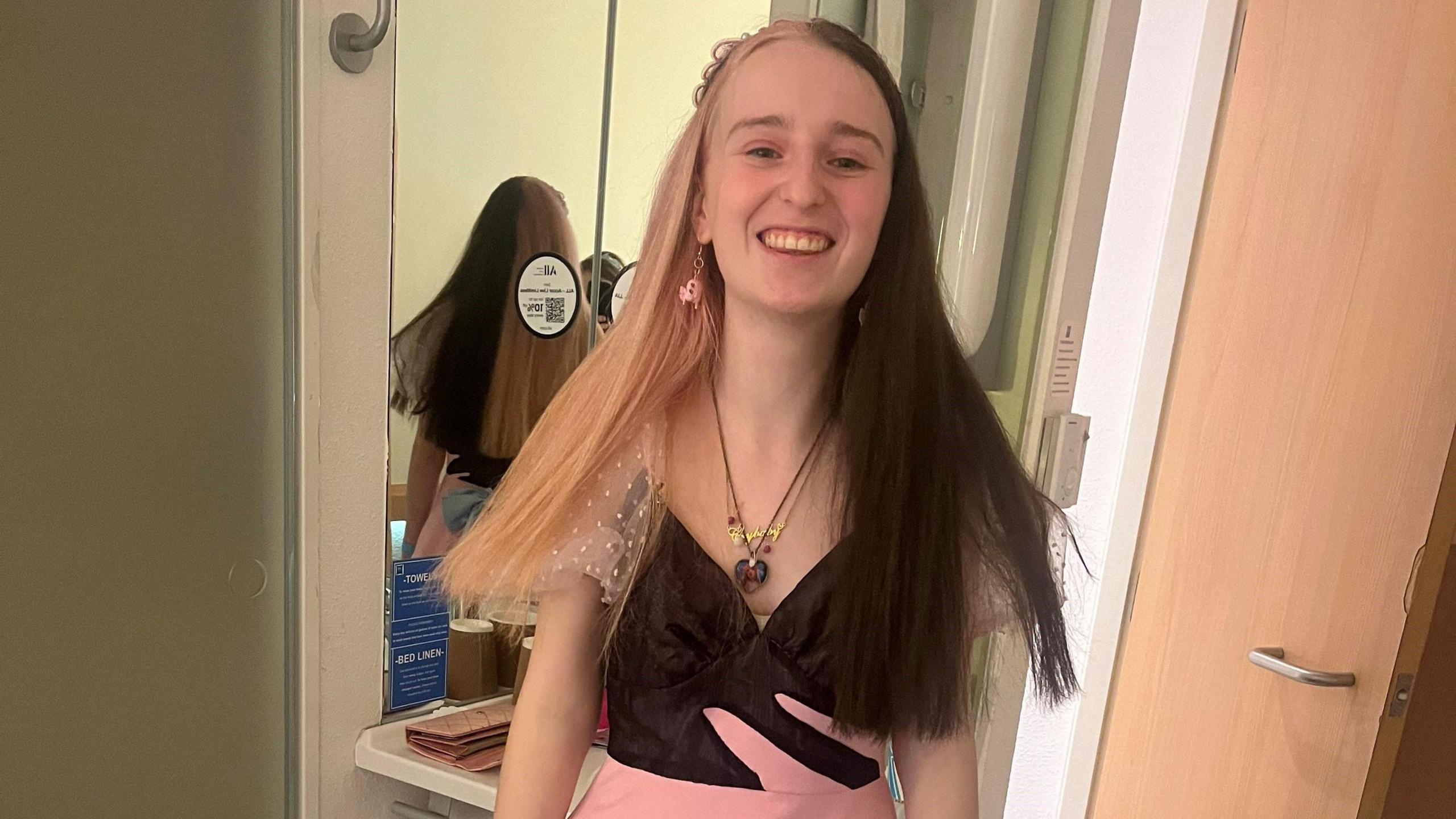 Lilly Hamer smiling at the camera and wearing a pink and black dress with short mesh sleeves. Her long hair is black on the left hand side from the middle parting down and blonde on the right hand side. She is wearing a heart-shaped necklace and pink pendant earrings.