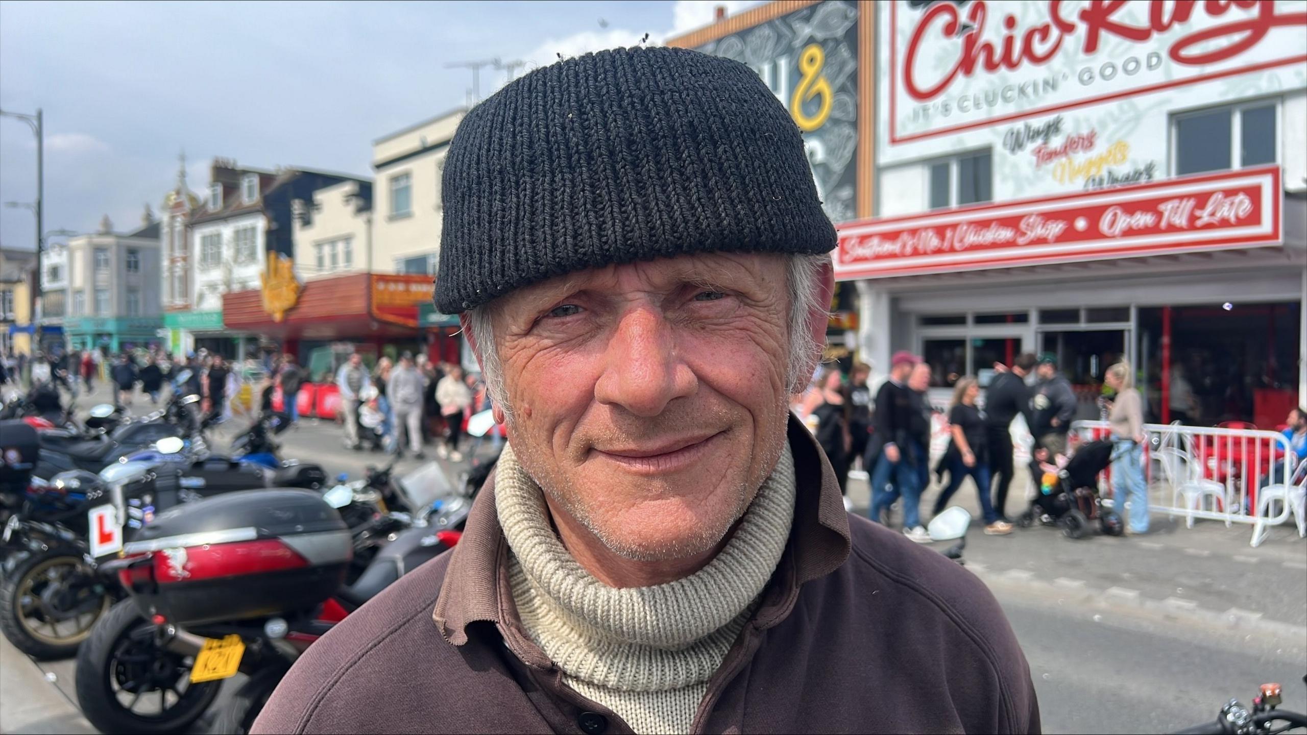 Mark Wilsmore smiles at the camera. He wears a knitted black beanie hat, a brown coat with a light green turtleneck jumper underneath. Parked motorbikes  and businesses along the Southend seafront can be seen behind him. 