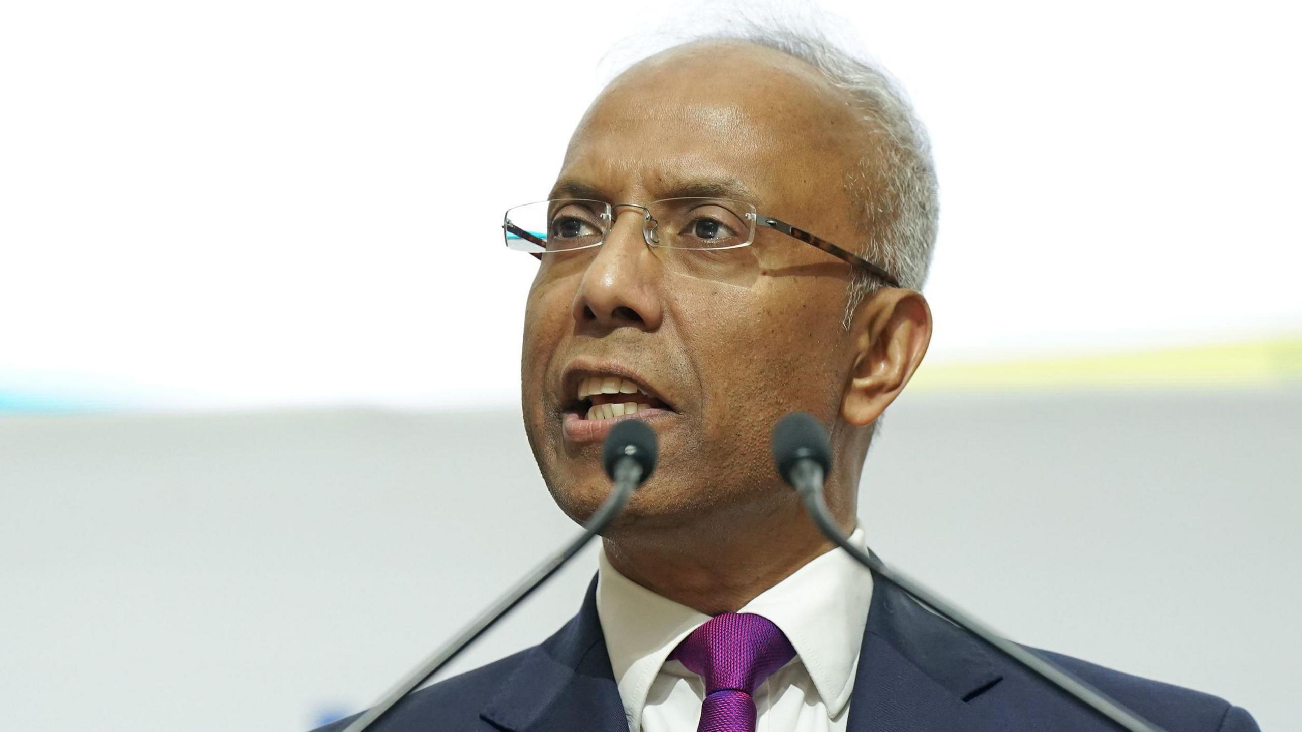 A medium close up of Lutfur Rahman speaking at an event. He is wearing glasses, wearing a navy suit with a white shirt and purple tie. 