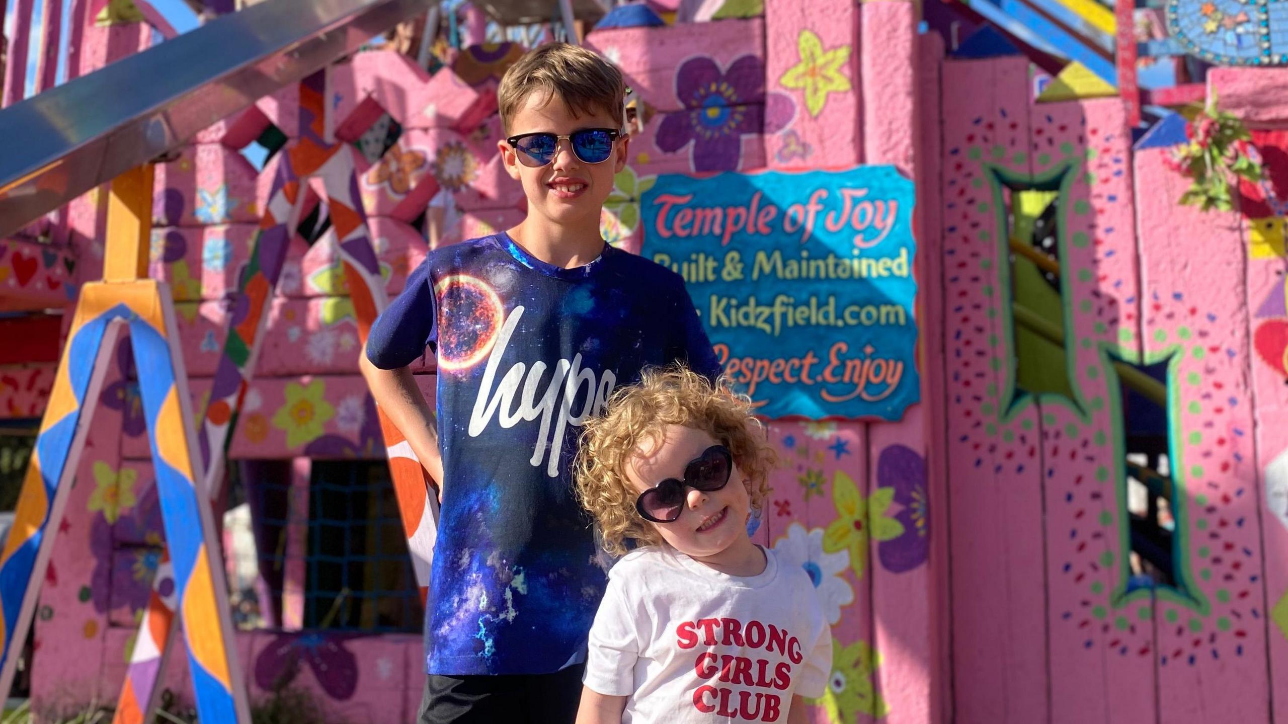 Kelly's children in front of a Temple of Joy sign, in the Kidzfield at the festival. Kelly's daughter is wearing a 'strong girls club' t shirt and her son is wearing a blue Hype t shirt. Both are wearing sunglasses.