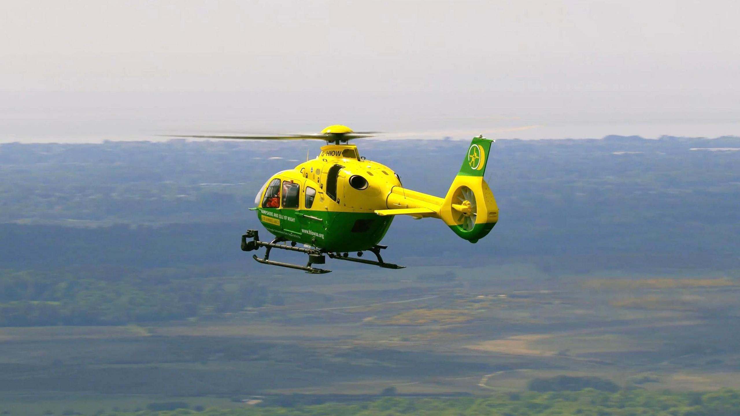 A green and yellow ambulance helicopter is flying mid-air against a backdrop of green land and the coast.
