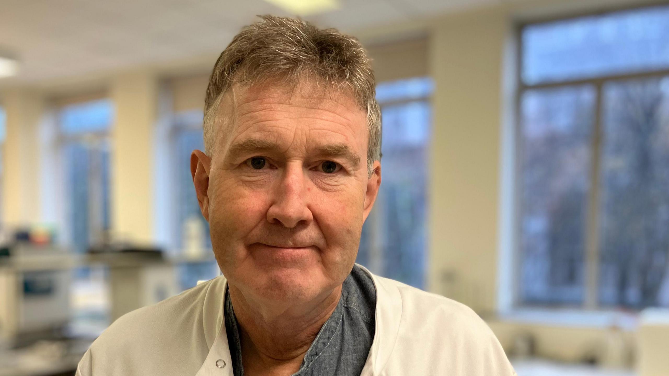 Prof Arwyn Tomos Jones looking at the camera. He is wearing a white lab coat over a blue shirt.