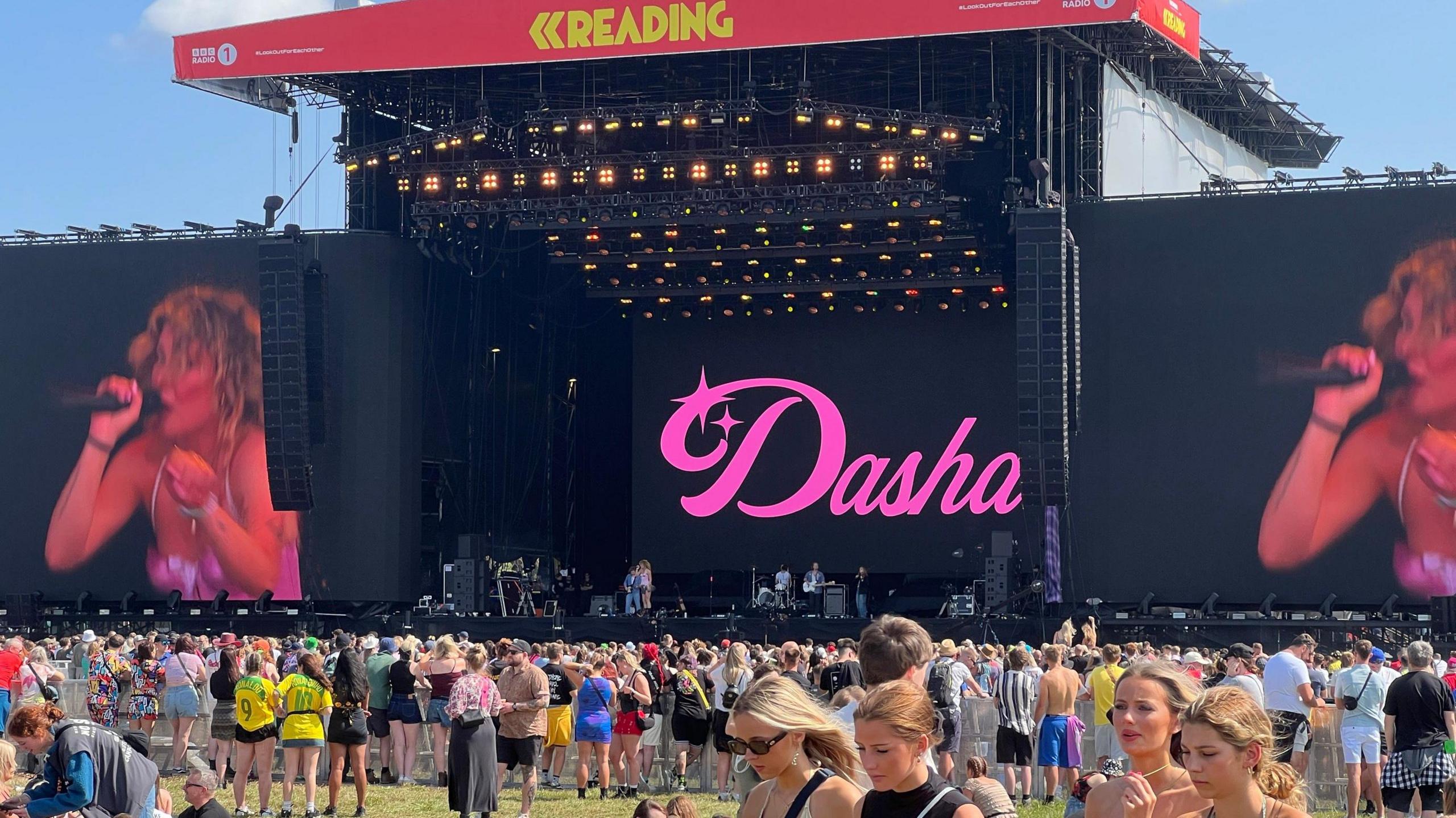 A crowd gathered in front of Reading Festival main stage where Dasha is performing. The pink Dasha logo is displayed at the back of the stage and she can be seen singing into a microphone on two screens on either side