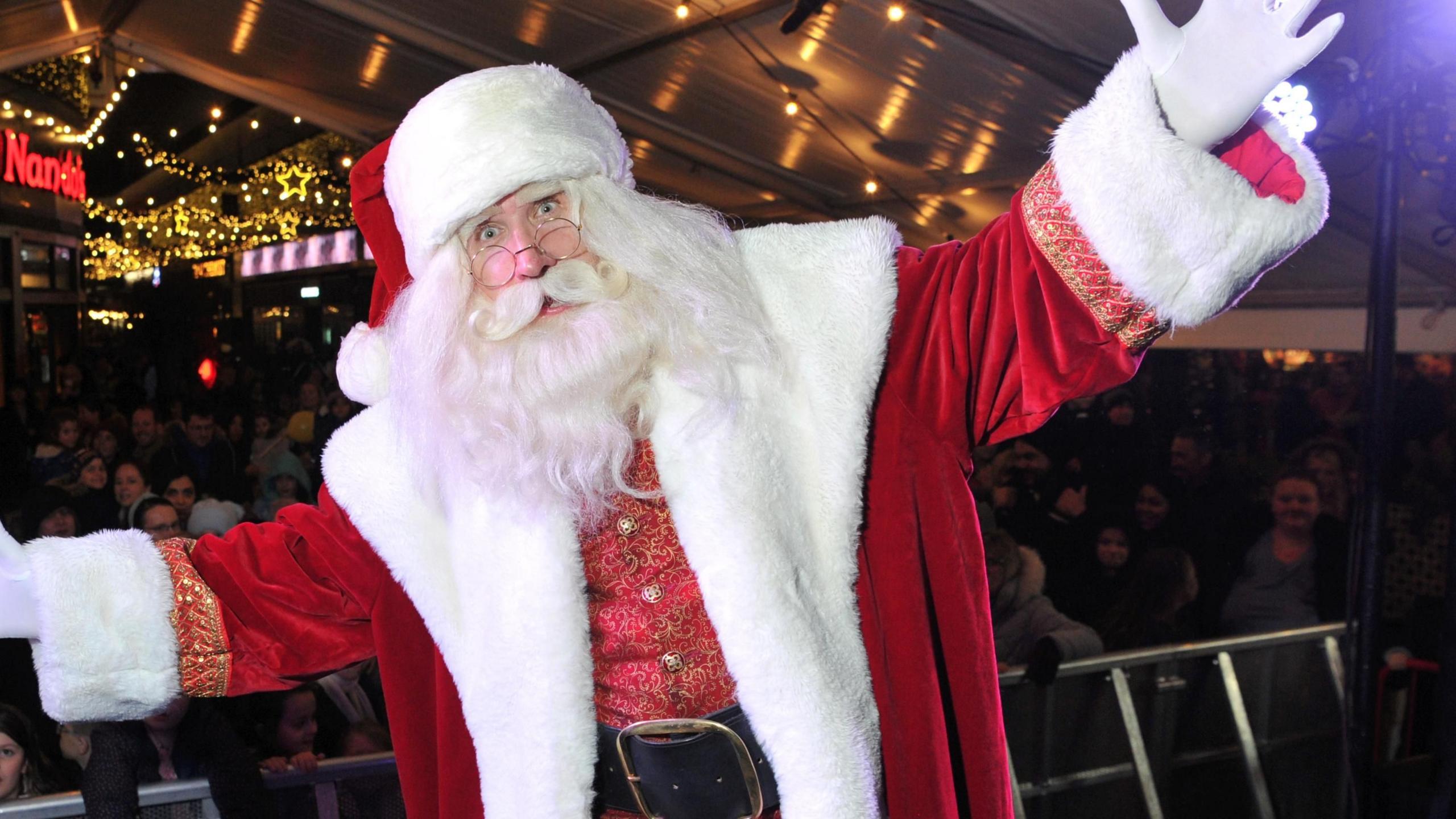 Santa Claus on stage in front of an audience posing with his arms stretched out. There is a Nando's and some Christmas lights behind him. 